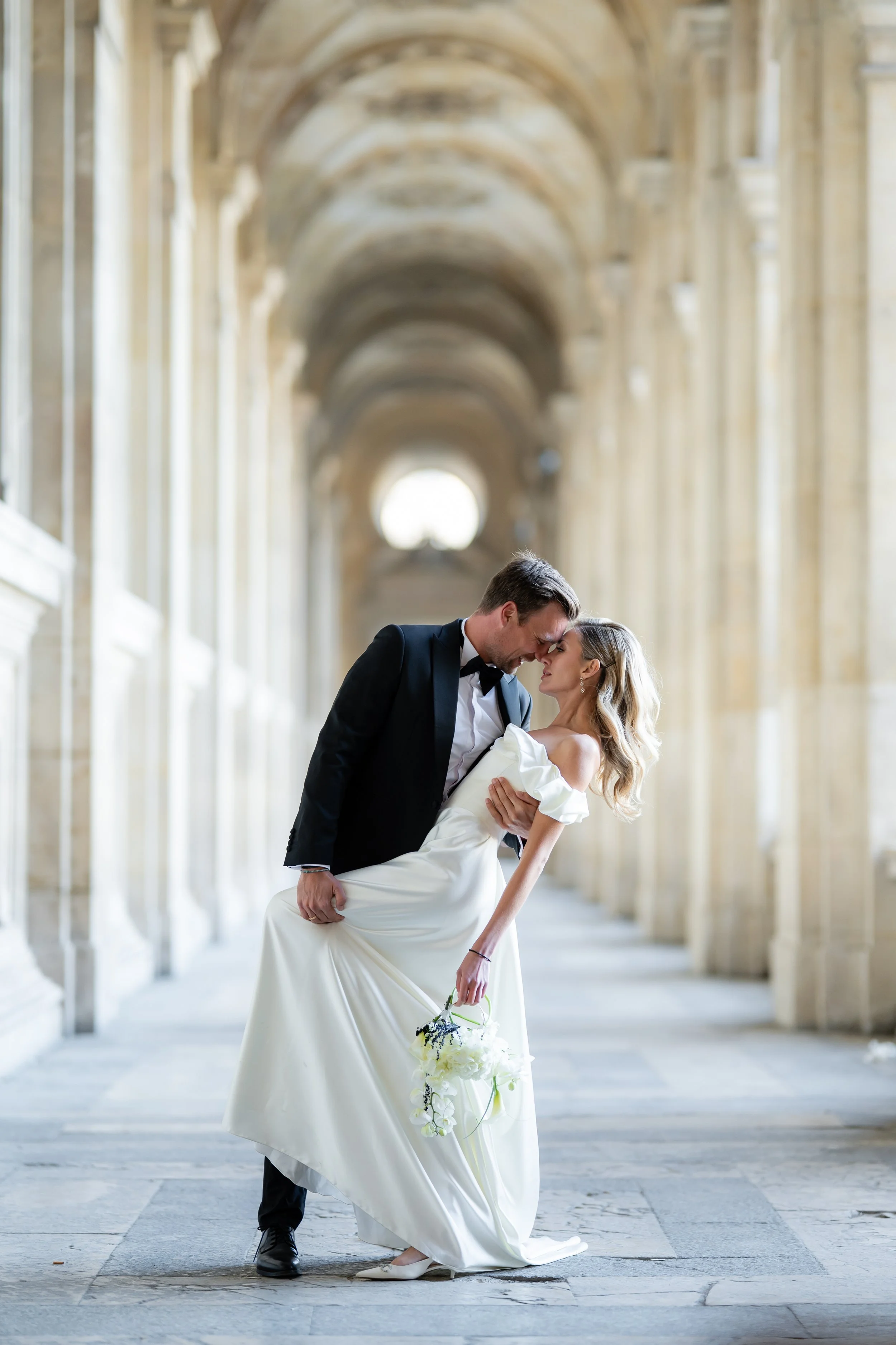A couple in wedding attire dancing in a grand, arched hallway with columns and vaulted ceilings at a wedding or special event.