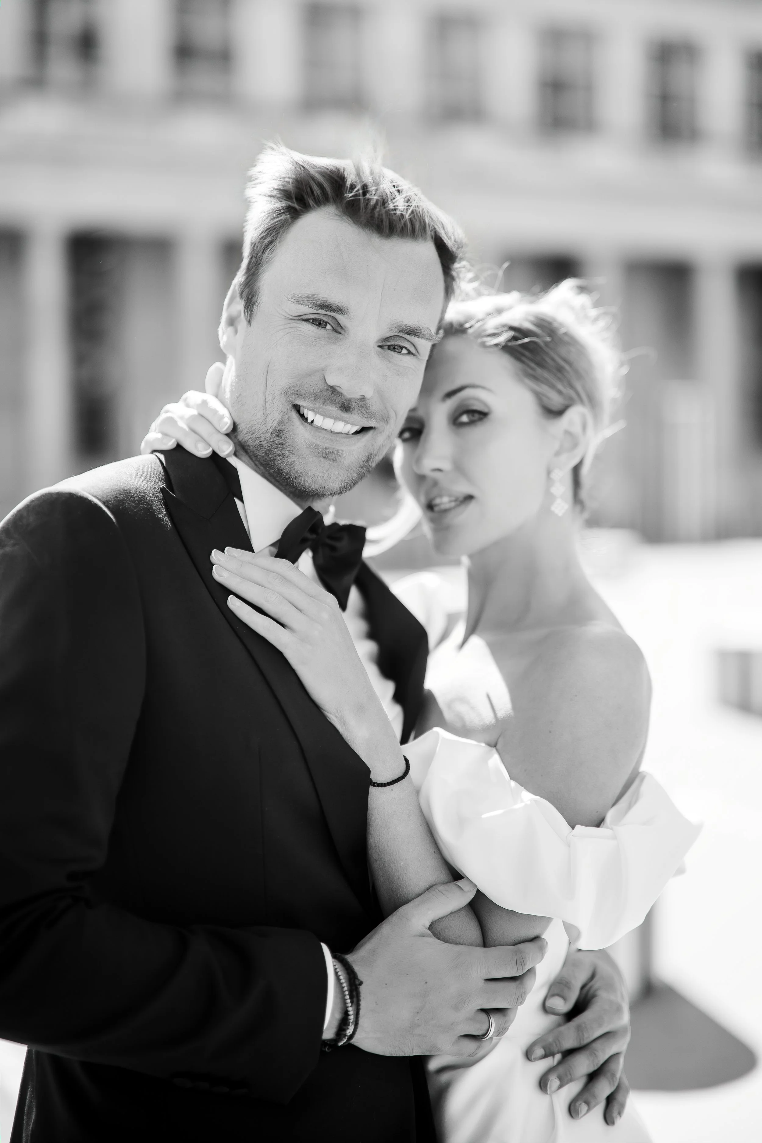 A smiling man in a tuxedo embraces a pregnant woman in a strapless white dress, both looking happy. They are outdoors with a blurred building in the background.