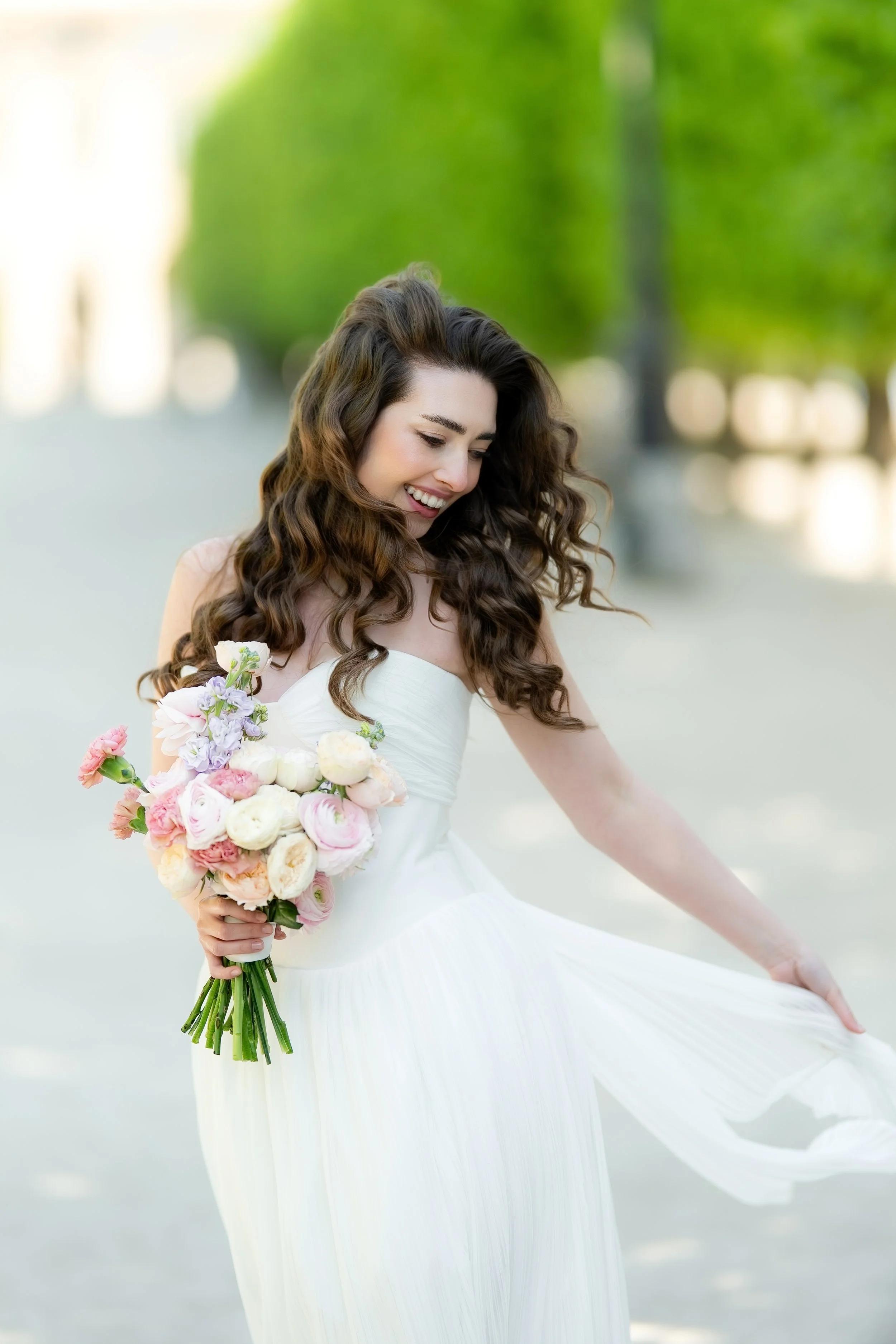 A smiling woman with wavy brown hair wearing a white dress, holding a bouquet of pastel-colored flowers, outdoors with blurry green trees in the background.