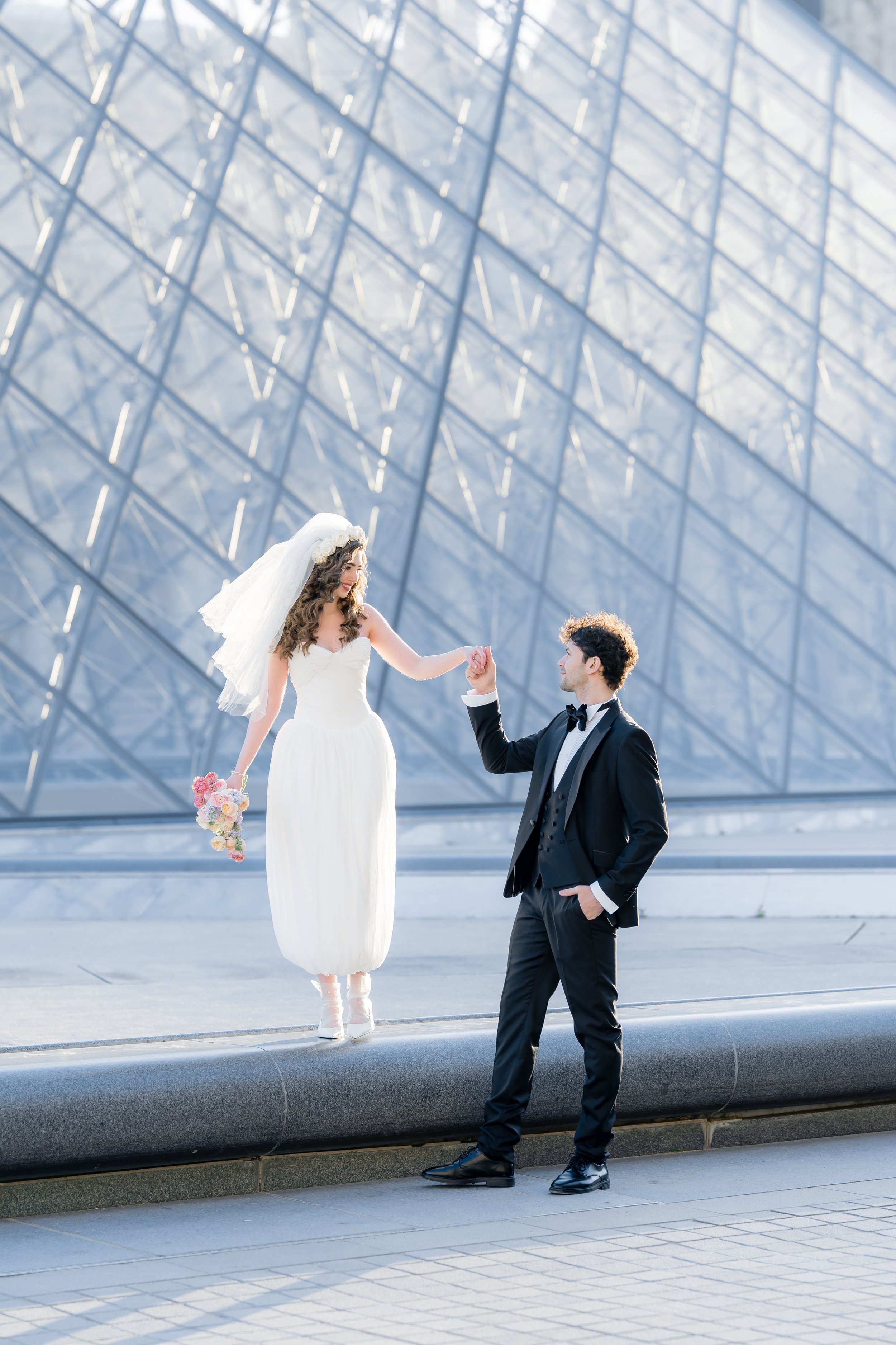 A bride in a white dress and veil holding a bouquet, standing on steps outside the louvre touching hands with a groom in a black tuxedo.