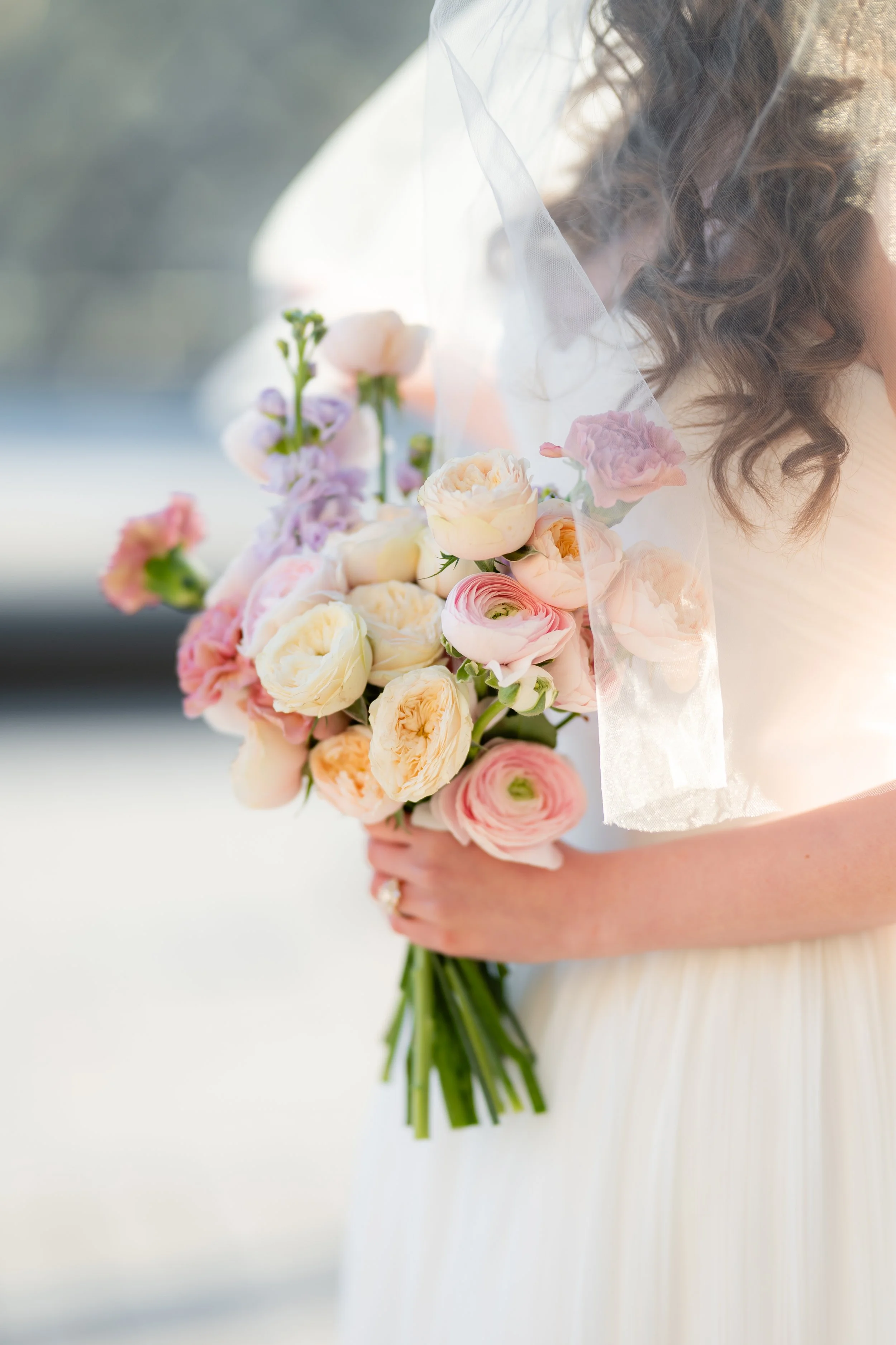 A bride holding a bouquet of pastel-colored roses and ranunculus flowers, with her face partially visible and her hair styled in loose curls, wearing a veil.
