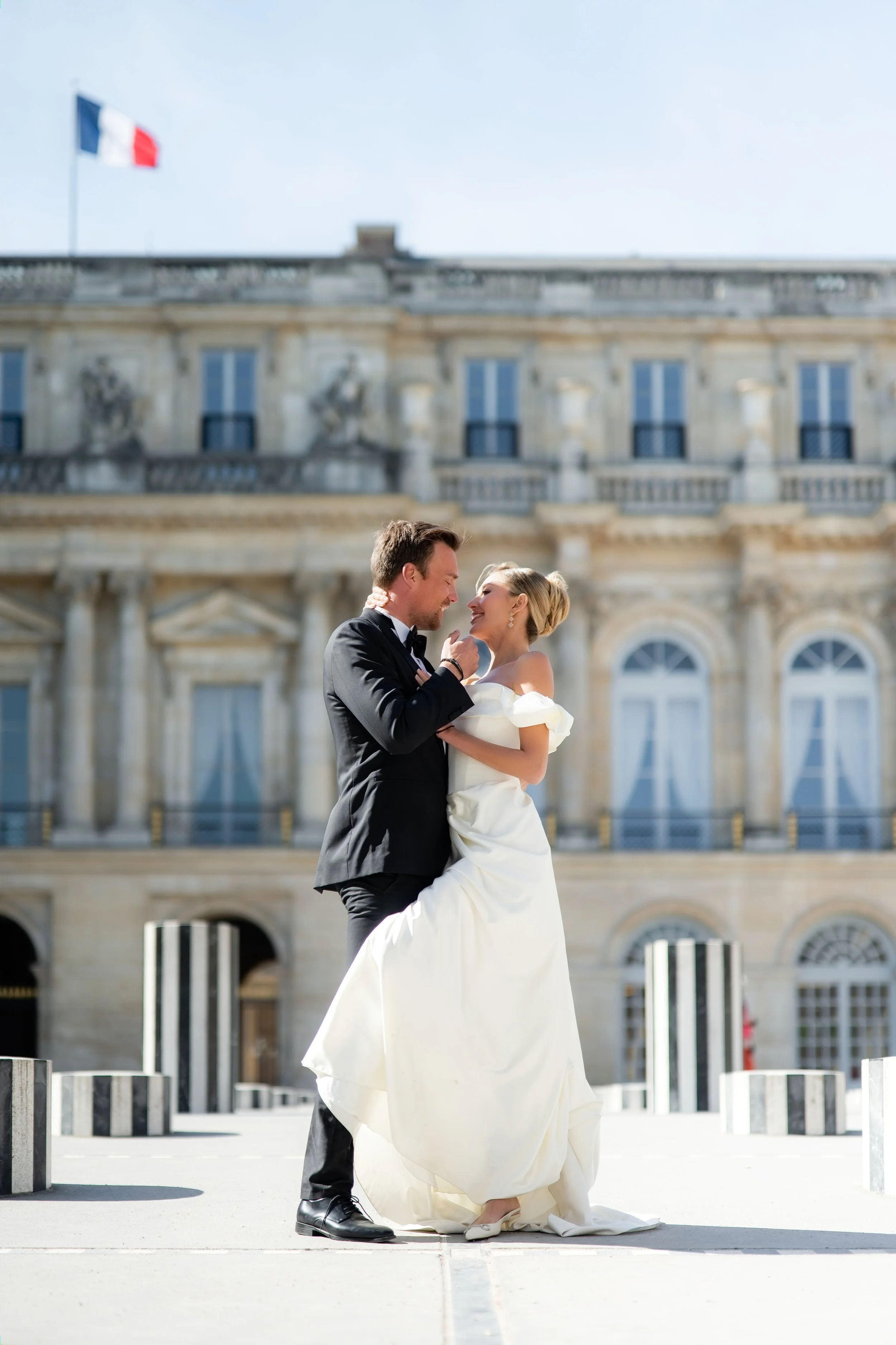 A groom and bride smiling and looking at each other outside a historic building with a French flag on top. The groom is in a black tuxedo and the bride is in an off-shoulder white wedding gown.