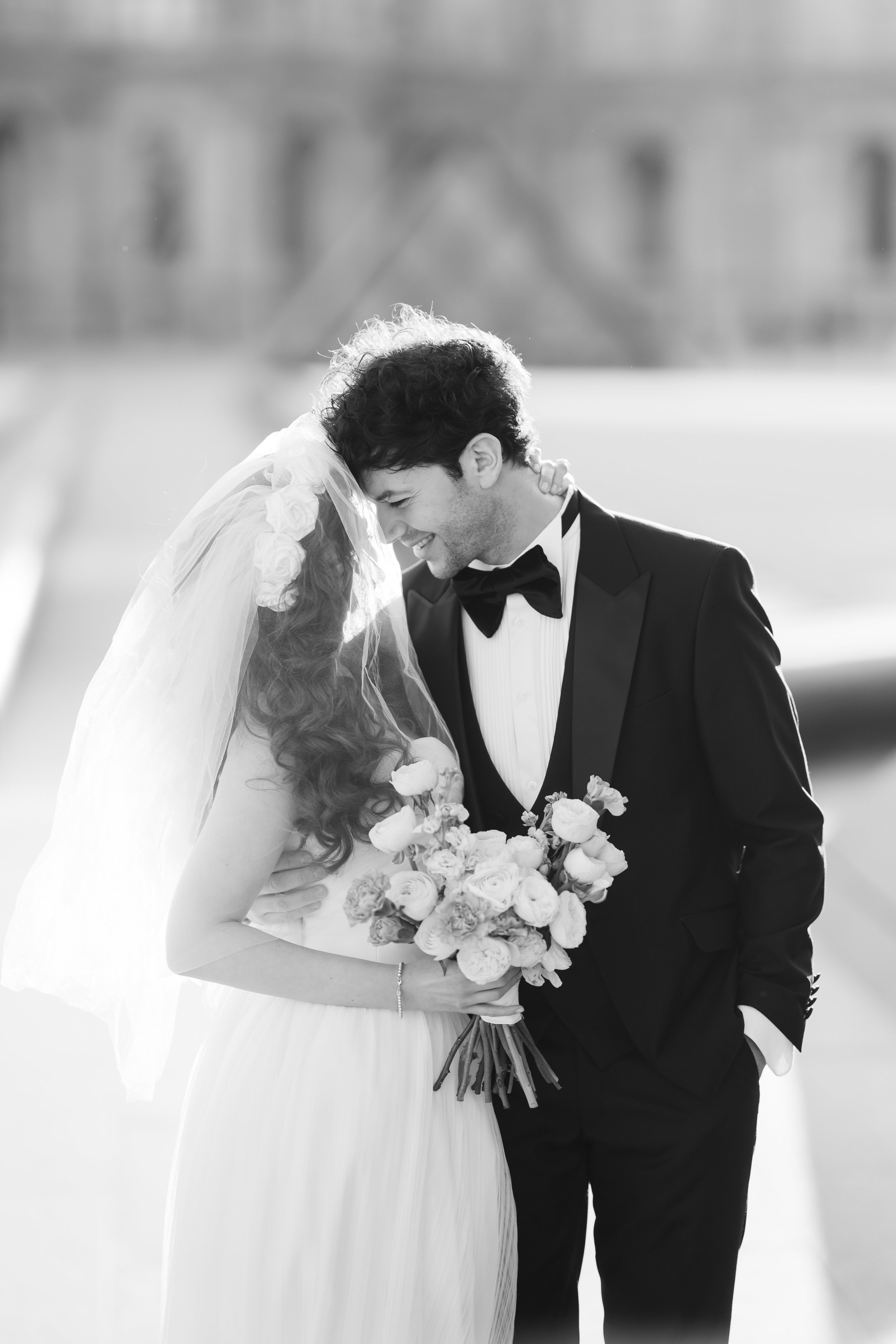 A bride and groom sharing a joyful moment outside, with the bride holding a bouquet of flowers. They are smiling and gently touching foreheads, dressed in wedding attire—she in a white gown with a veil decorated with roses, and he in a tuxedo with a 