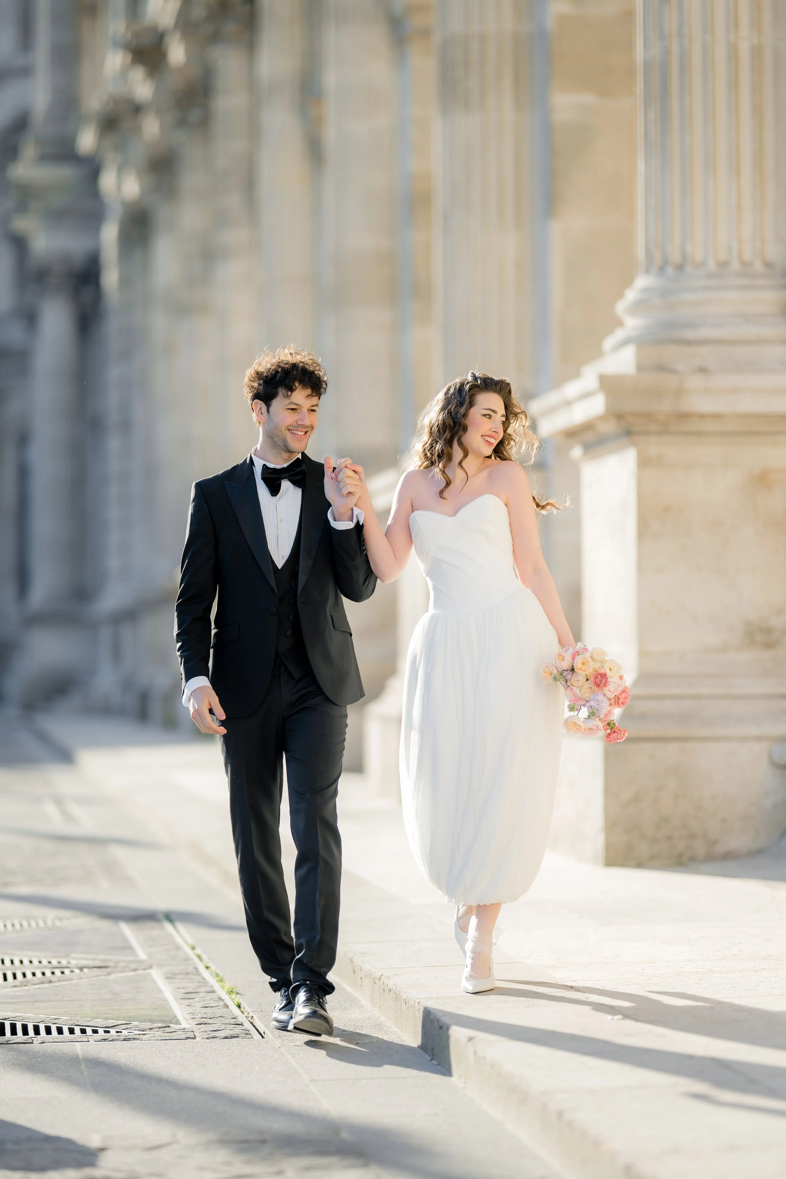 A smiling young couple, dressed in wedding attire, walking hand-in-hand outdoors with classical columns and a historic building in the background, during daytime.