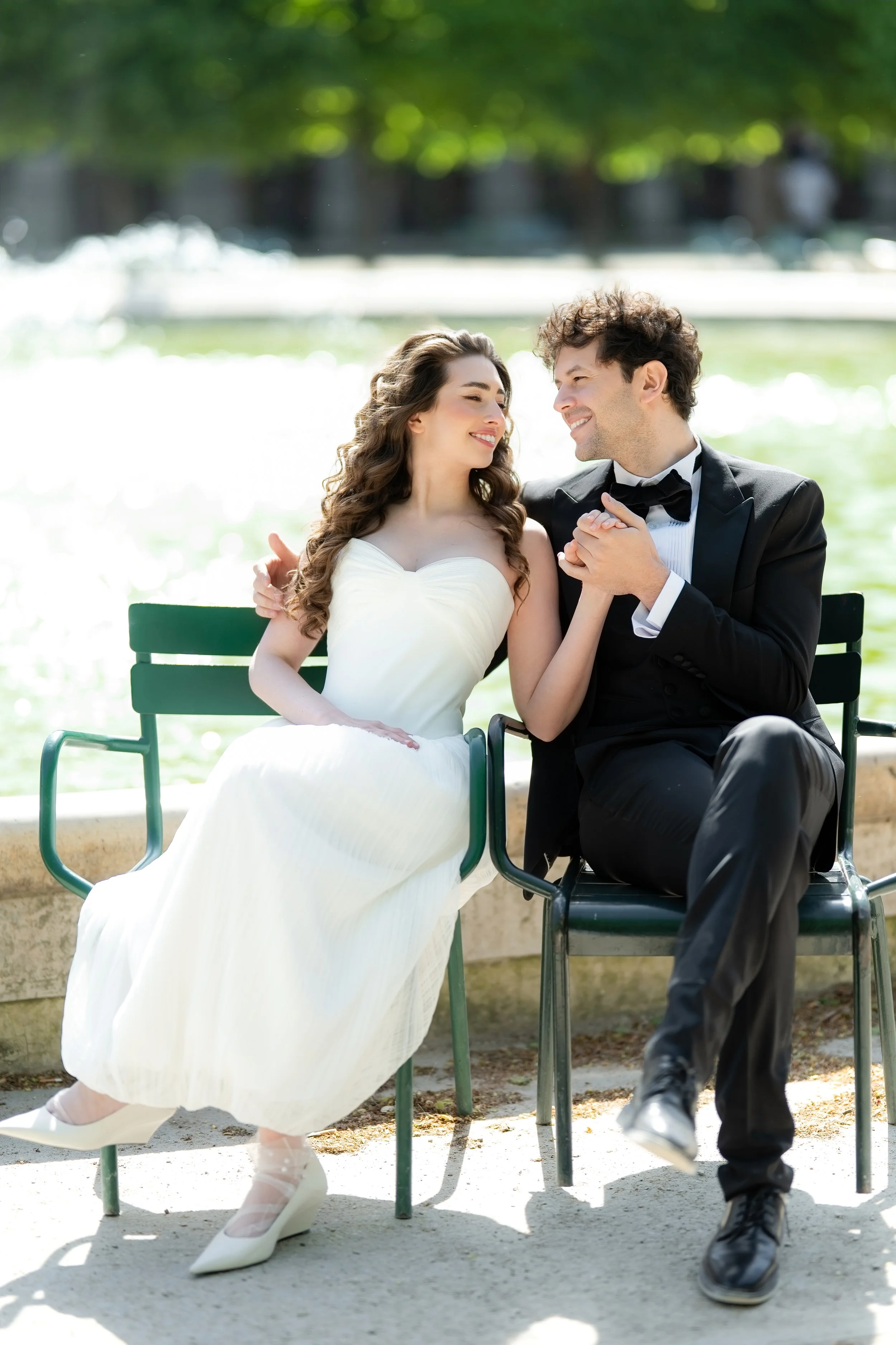 A bride and groom sitting close together on a park bench, smiling and looking at each other, with sunlight filtering through trees in the background.