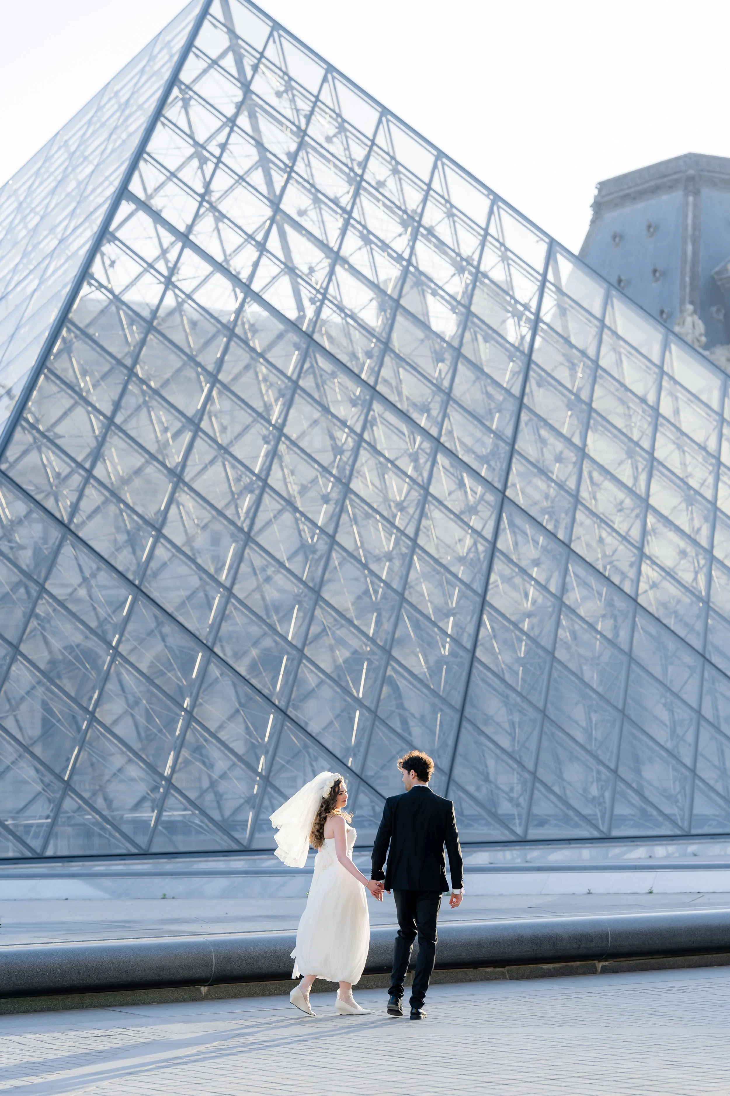 A bride and groom holding hands, walking in front of the Louvre building in daylight. The bride is wearing a white wedding dress and veil, and the groom is wearing a black suit. The couple is seen from behind, with the bride looking at the groom.
