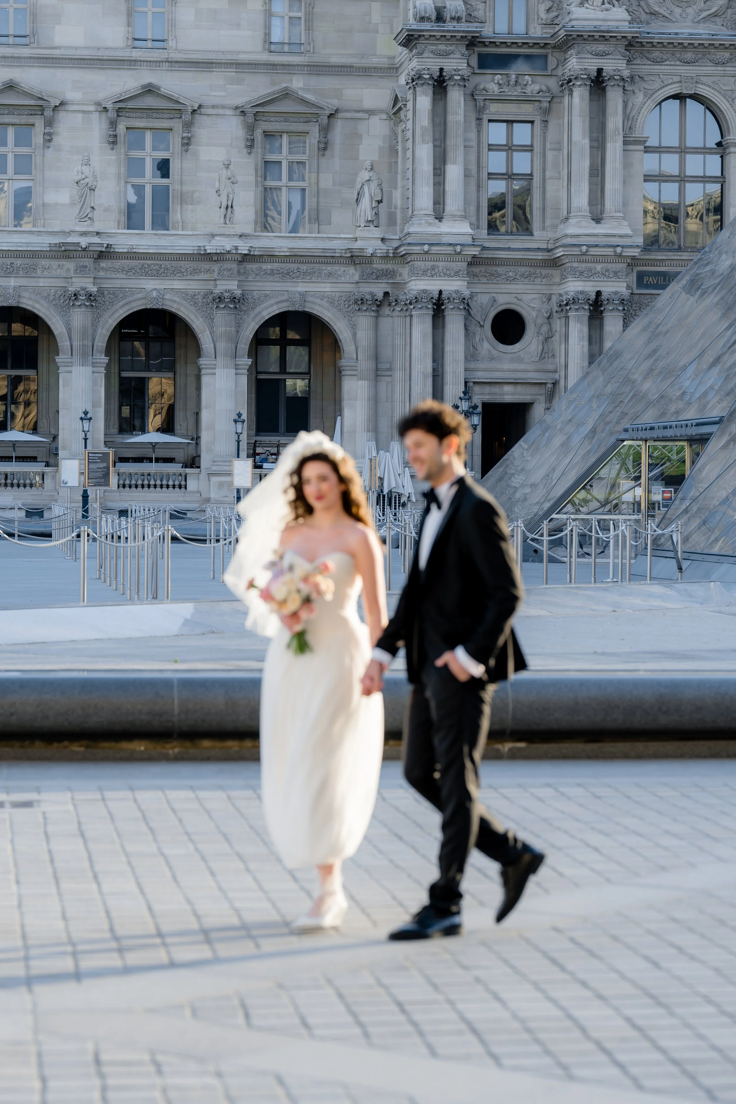 Blurred bride and groom walking hand in hand in front of historic building with statues and arched windows.