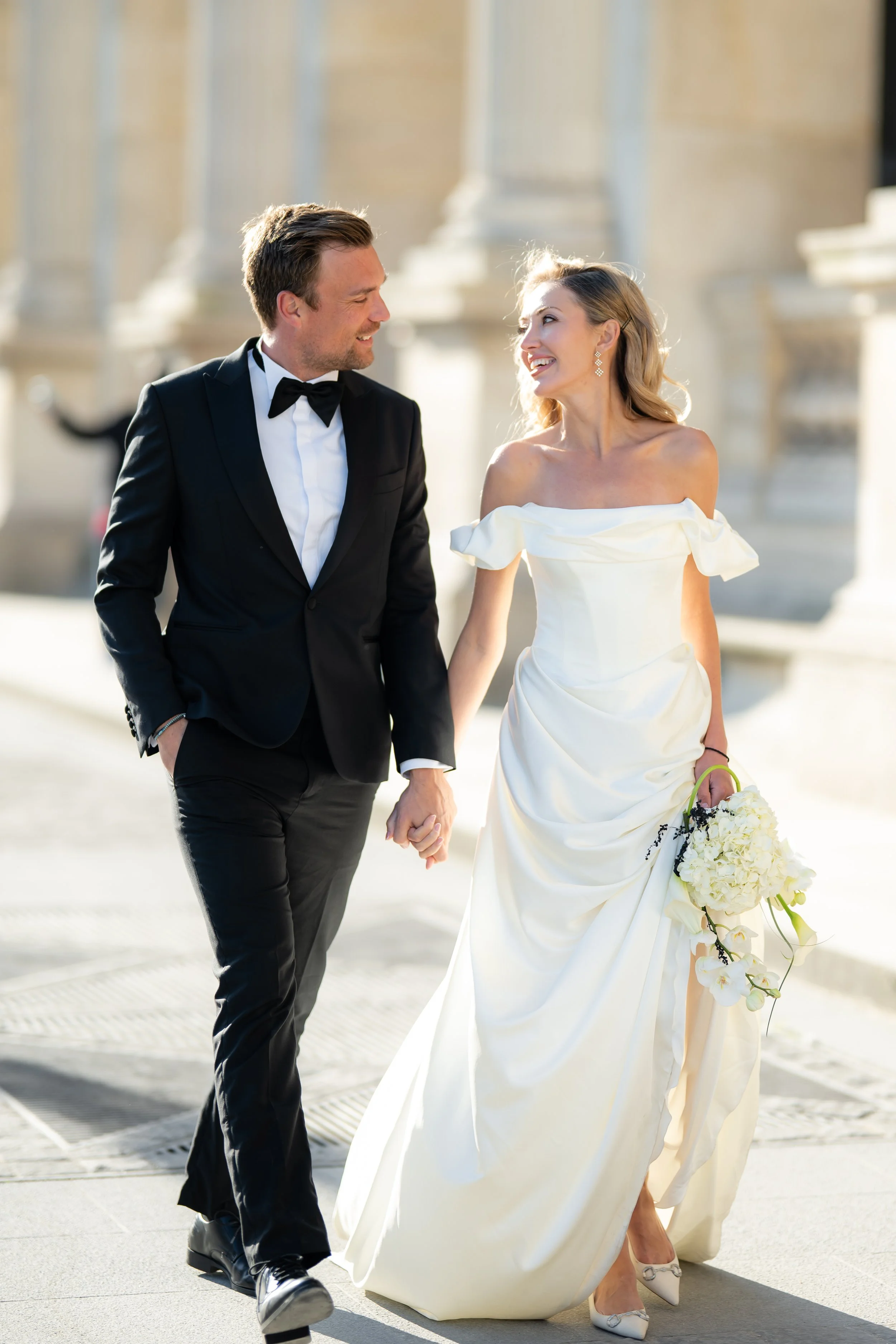 A man and woman, dressed in formal wedding attire, holding hands and looking at each other while walking outdoors.
