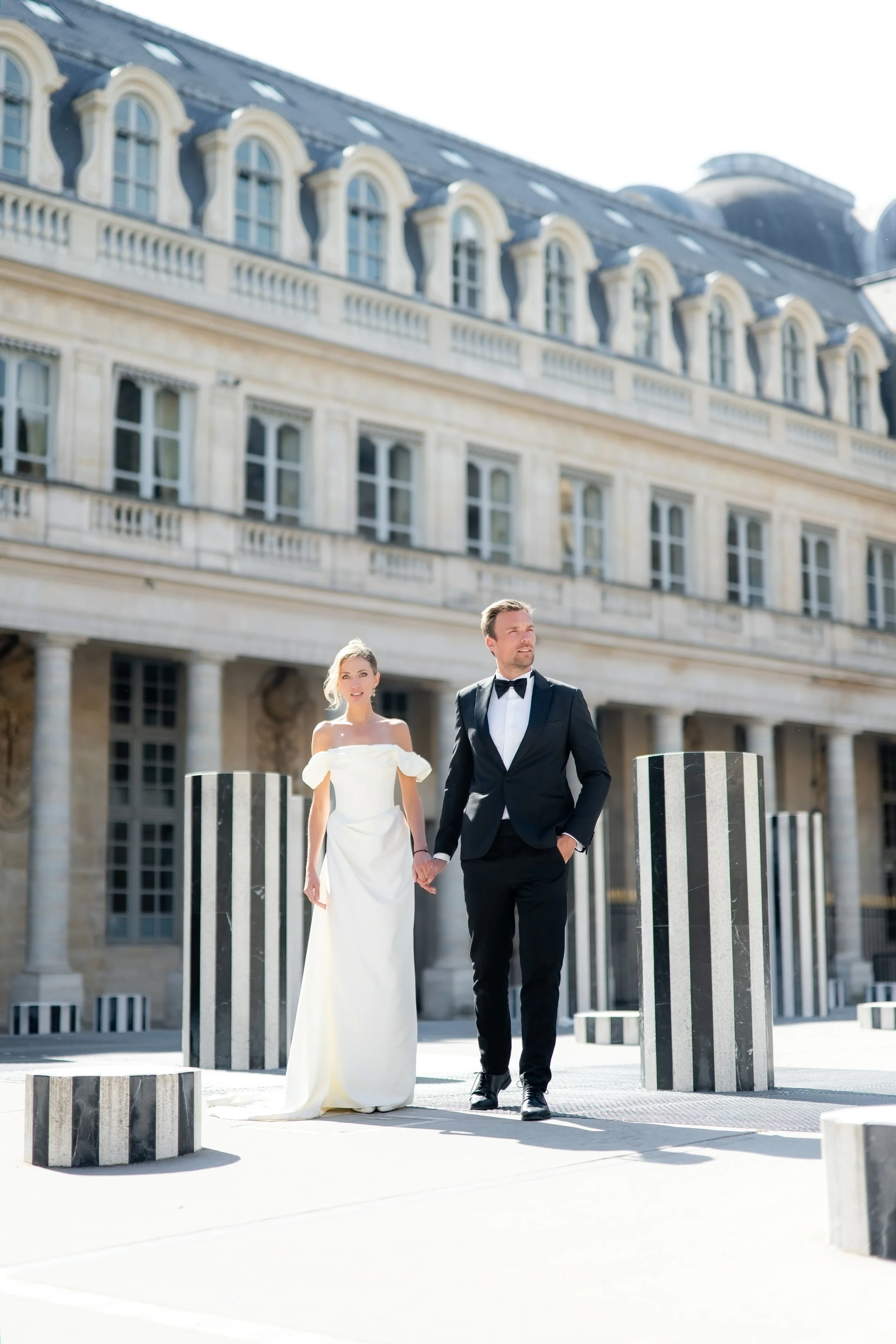A bride and groom holding hands walking outdoors in front of an elegant historic building with arched windows and a mansard roof, on a sunny day.