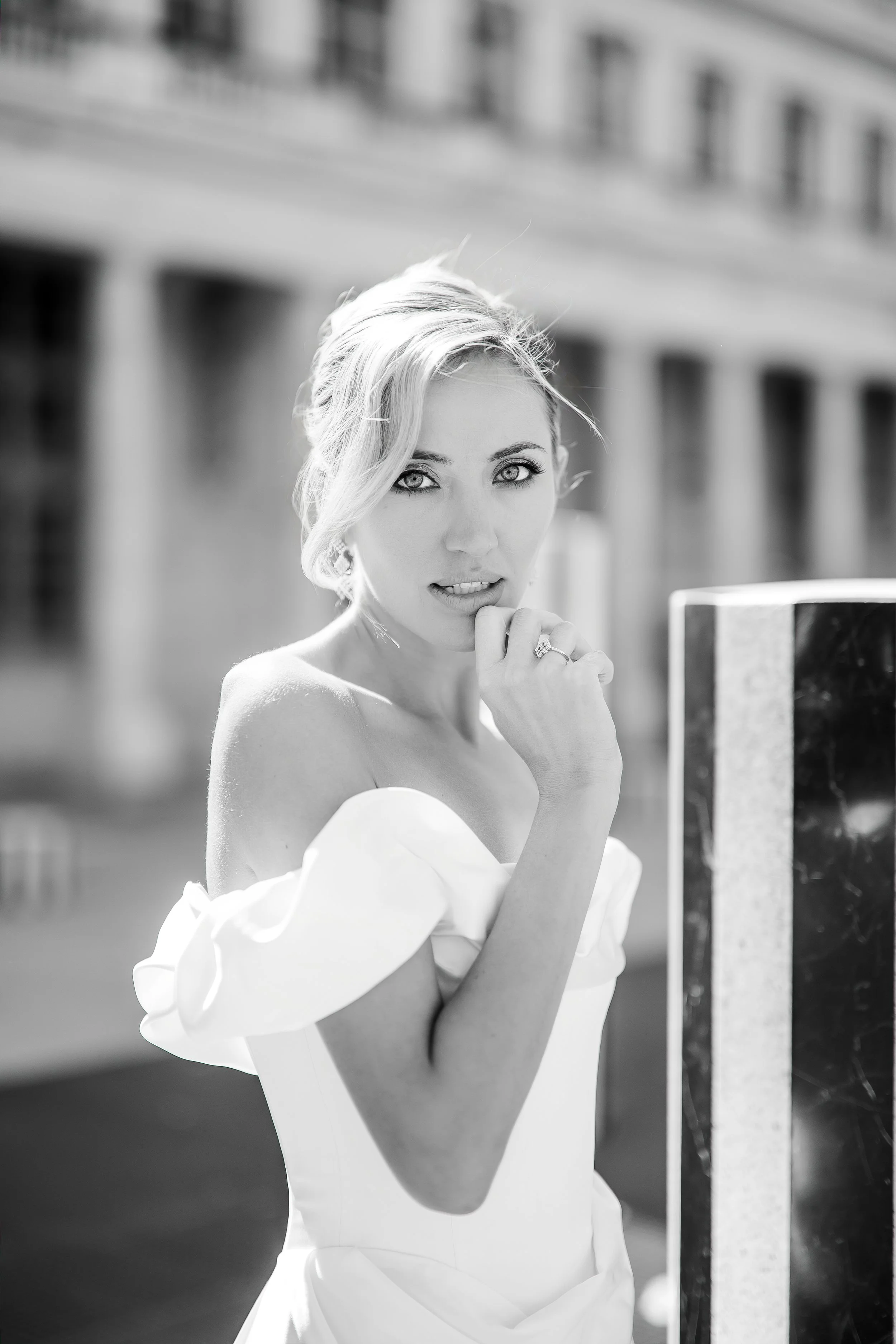 Black and white photo of a woman with short, wavy blonde hair, looking at the camera with her hand near her face, outdoors in a city environment with a building in the background.