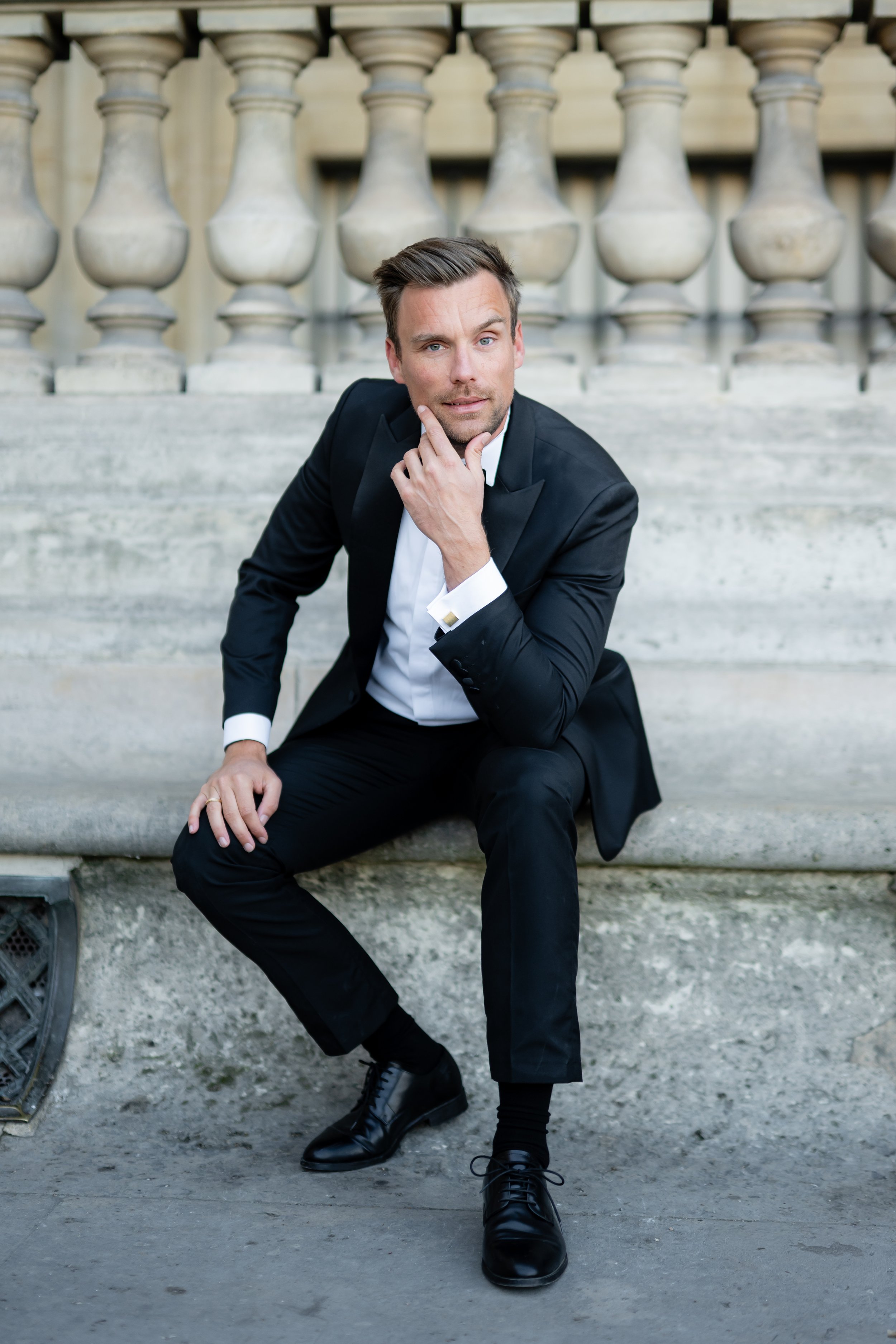 A man in a black tuxedo sitting on stone steps in front of a stone balustrade, with his hand on his chin, posing thoughtfully.