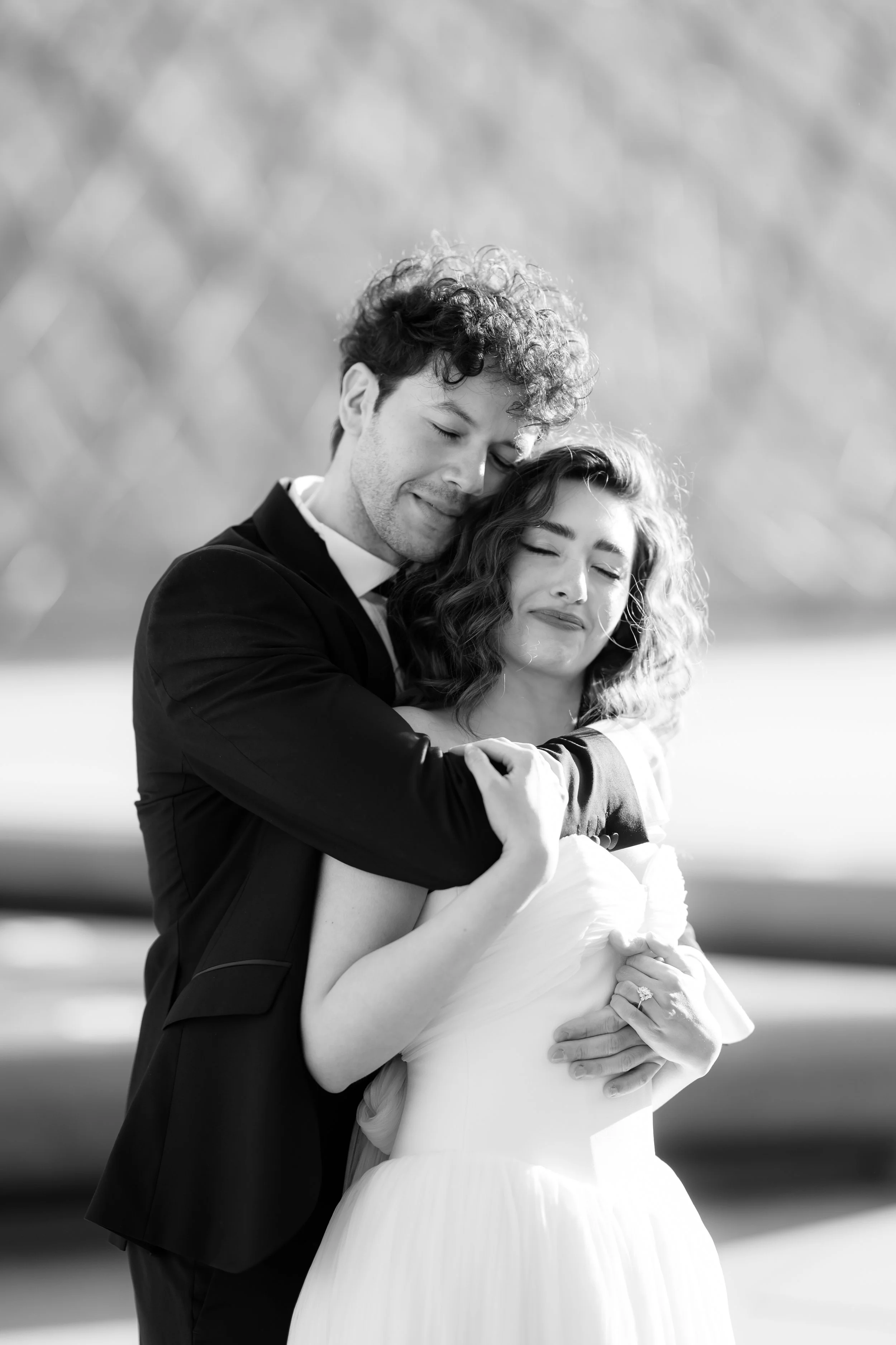 Black and white photo of a couple embracing outdoors. The man has curly hair and is wearing a tuxedo, and the woman has wavy hair and is wearing a wedding dress. They both have closed eyes and expressions of contentment.