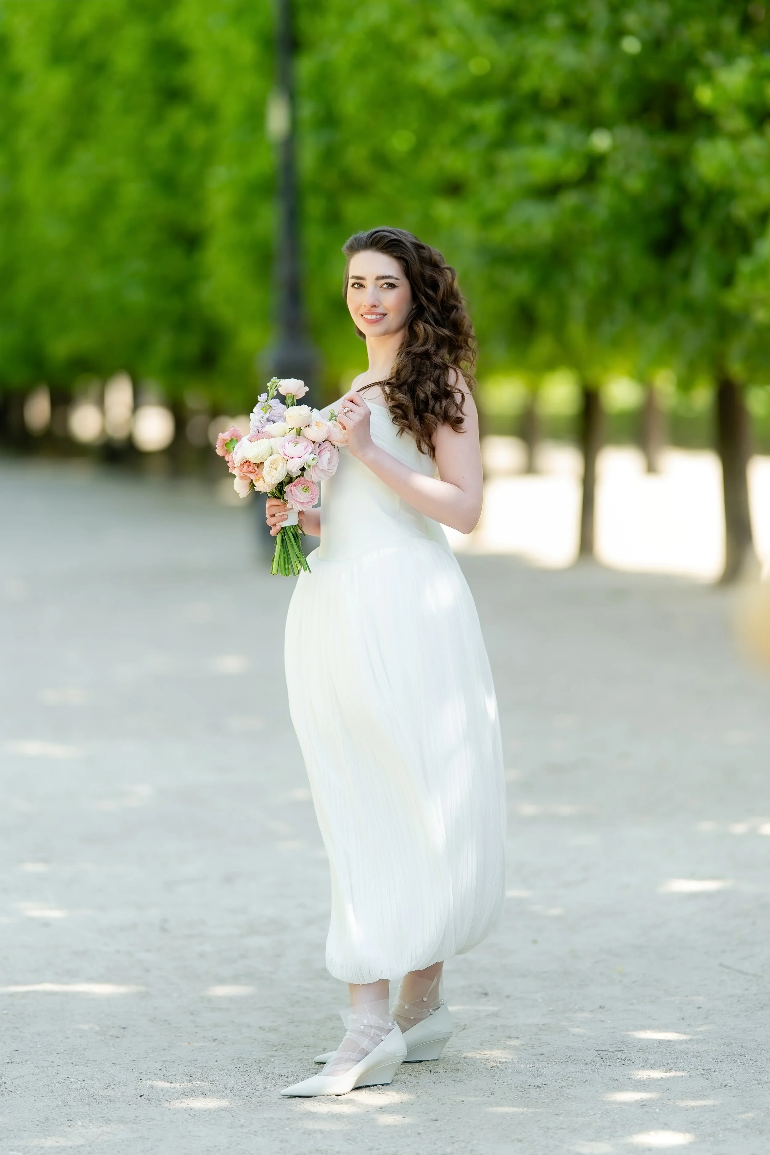 A woman in a white dress is holding a bouquet of pink and white flowers, standing outdoors on a sunny day with green trees in the background.