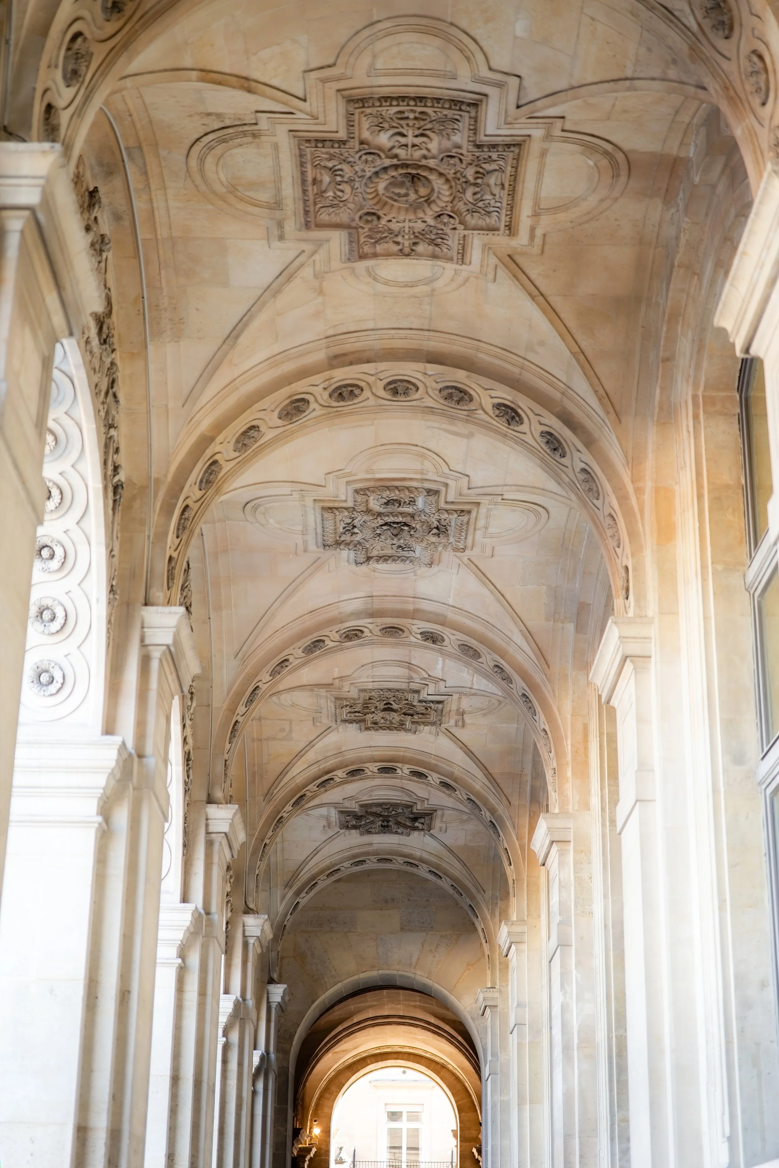 A grand hallway with high arched ceilings decorated with ornate carvings and moldings, featuring a series of architectural arches with intricate details, illuminated by natural light from windows and a warm glow from lamps at the far end.
