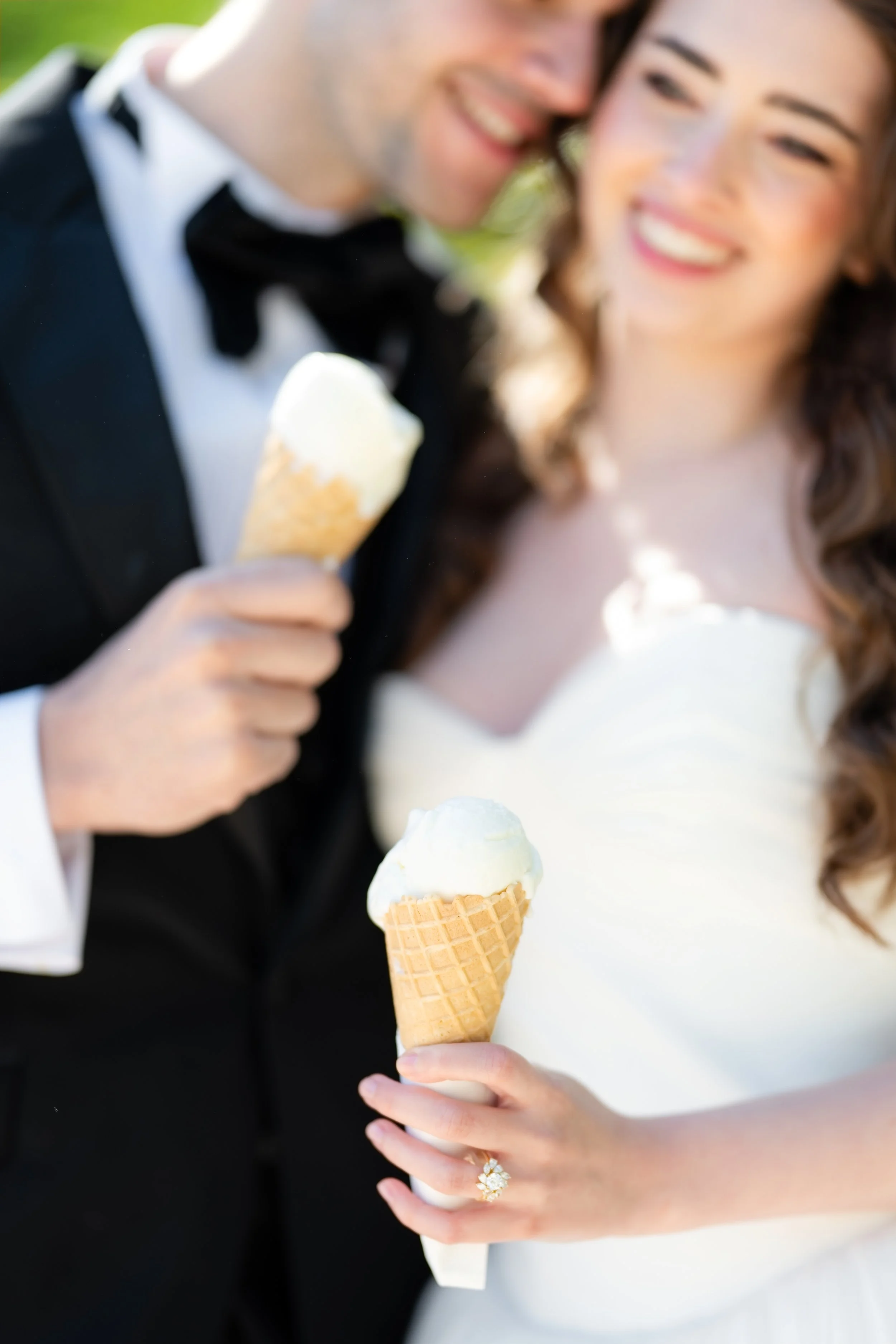 Close-up of a smiling couple, the groom in a black tuxedo with a bow tie and the bride in a white dress, both holding ice cream cones.