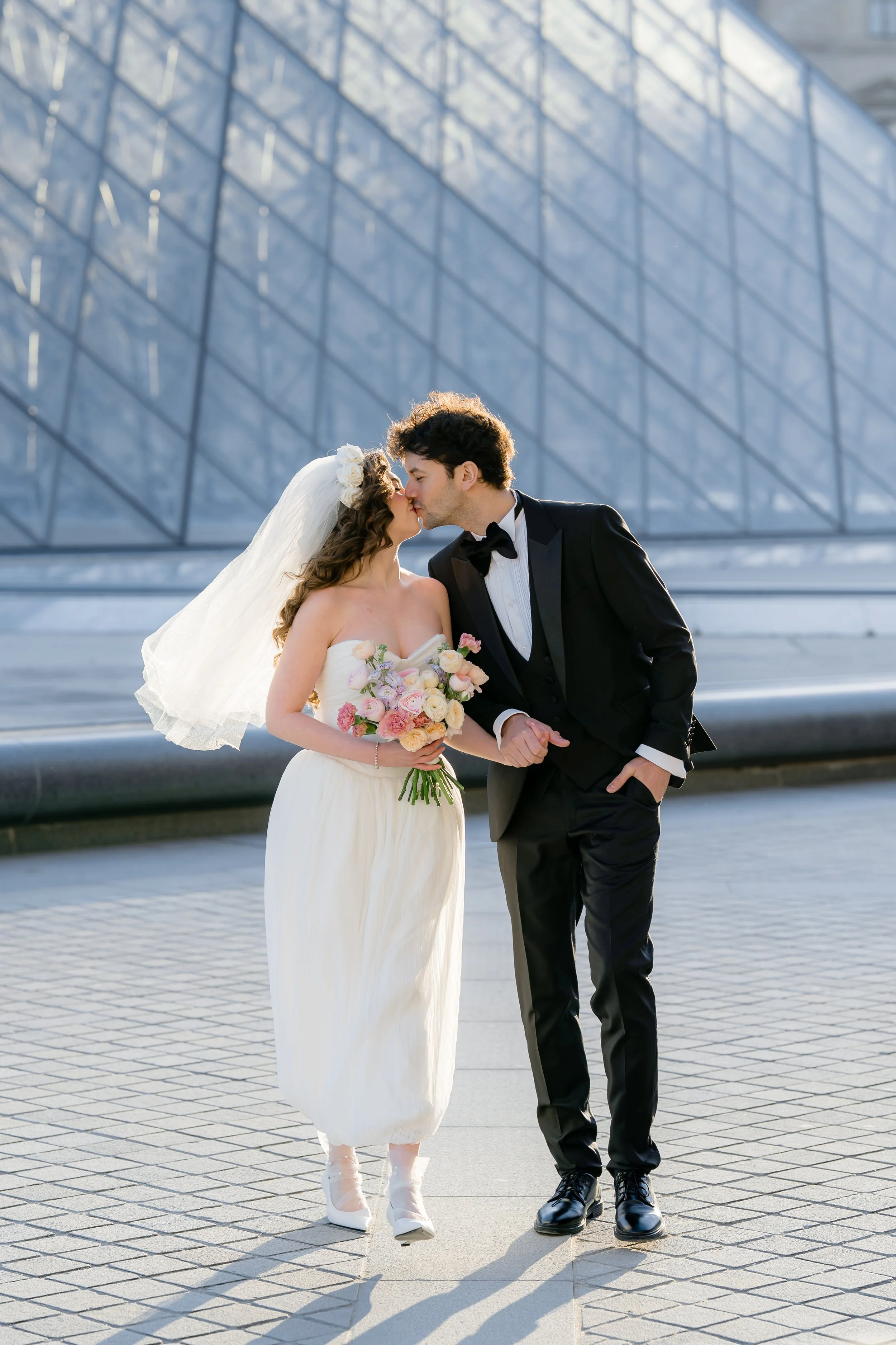 A bride and groom kissing outdoors in front of a modern glass building. The bride is wearing a white strapless wedding gown with a veil and holding a bouquet of pink and white flowers. The groom is in a black tuxedo with a bow tie.