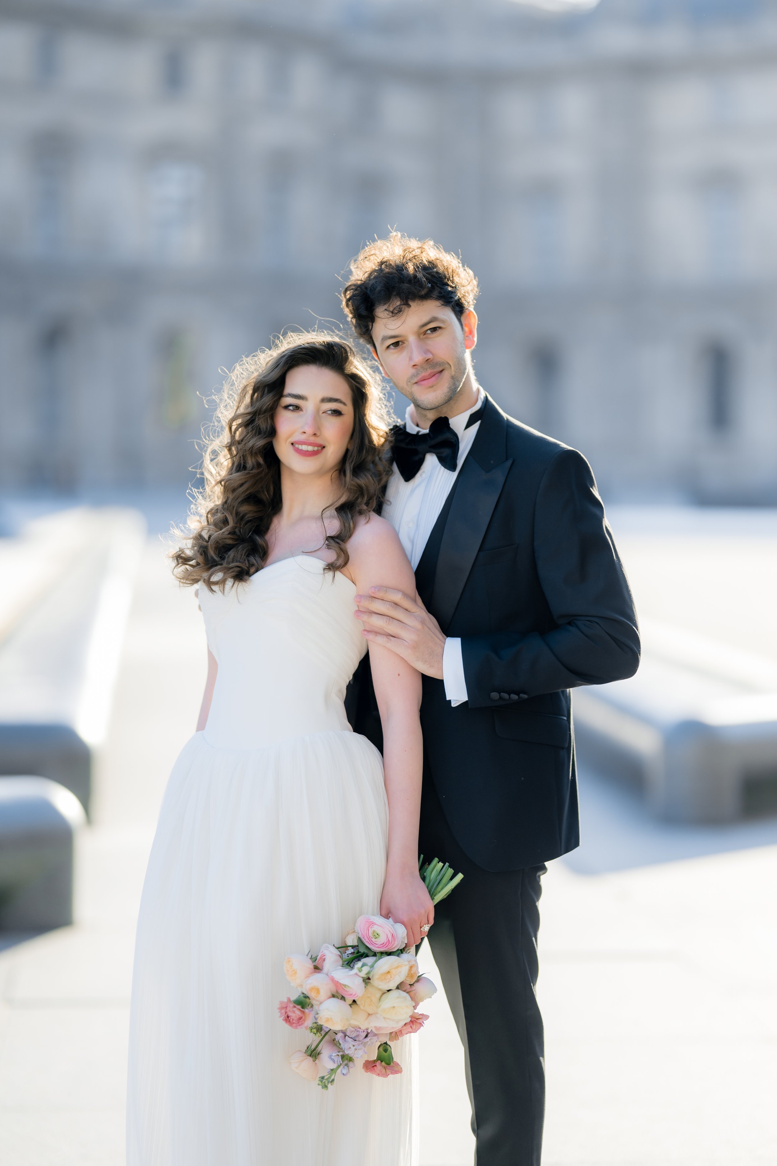 A young couple dressed in wedding attire, standing outdoors with an elegant building in the background. The woman wears a white wedding dress and holds a bouquet of pink and white flowers, while the man wears a black tuxedo and bow tie. Both are smil