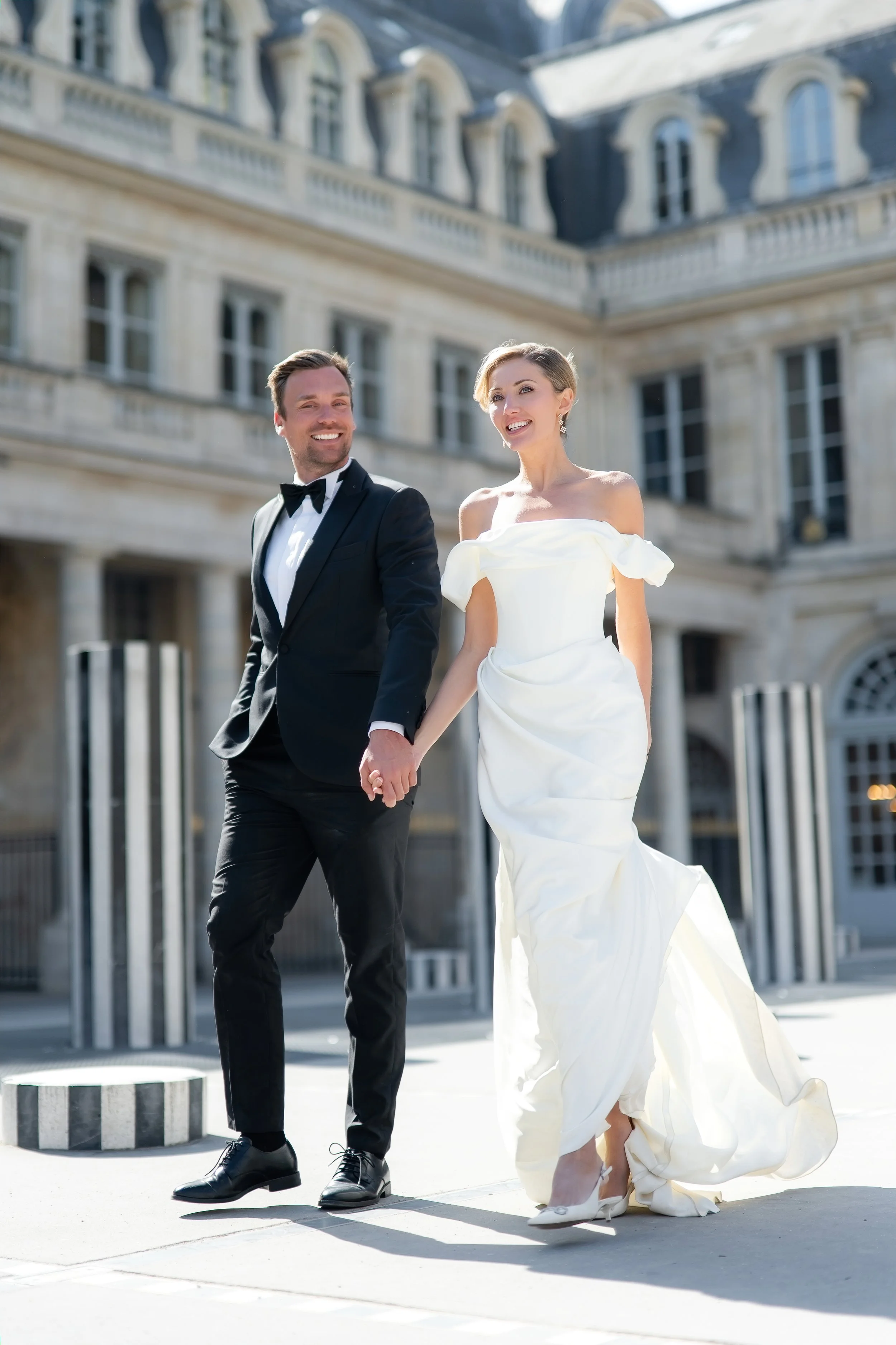 A happy couple dressed in wedding attire walking hand in hand outside a grand building with large windows and decorative architecture, both smiling and looking away from the camera.