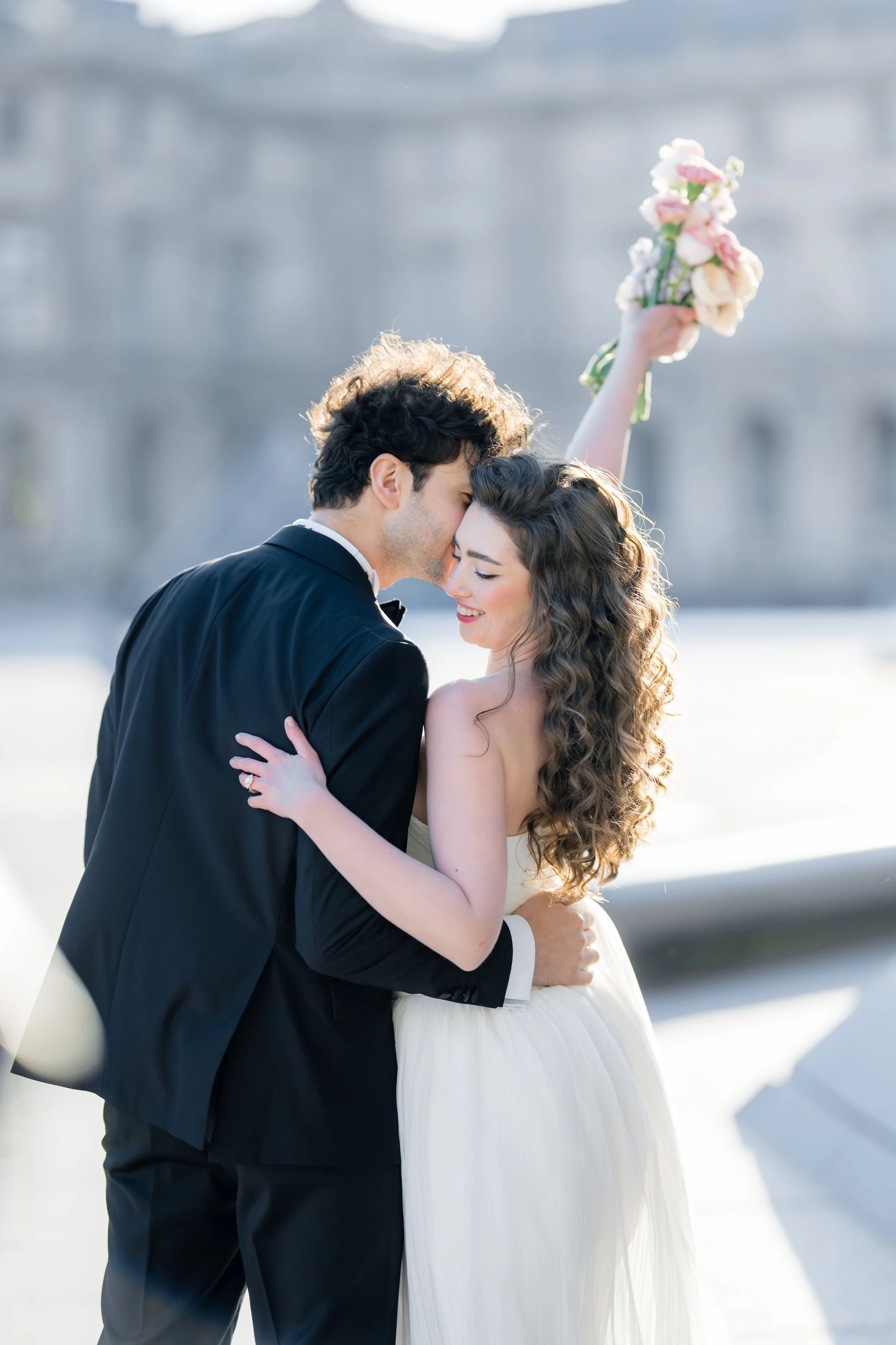 A bride and groom share a romantic moment outdoors, with the groom in a tuxedo and the bride in a wedding gown, holding a bouquet of flowers.