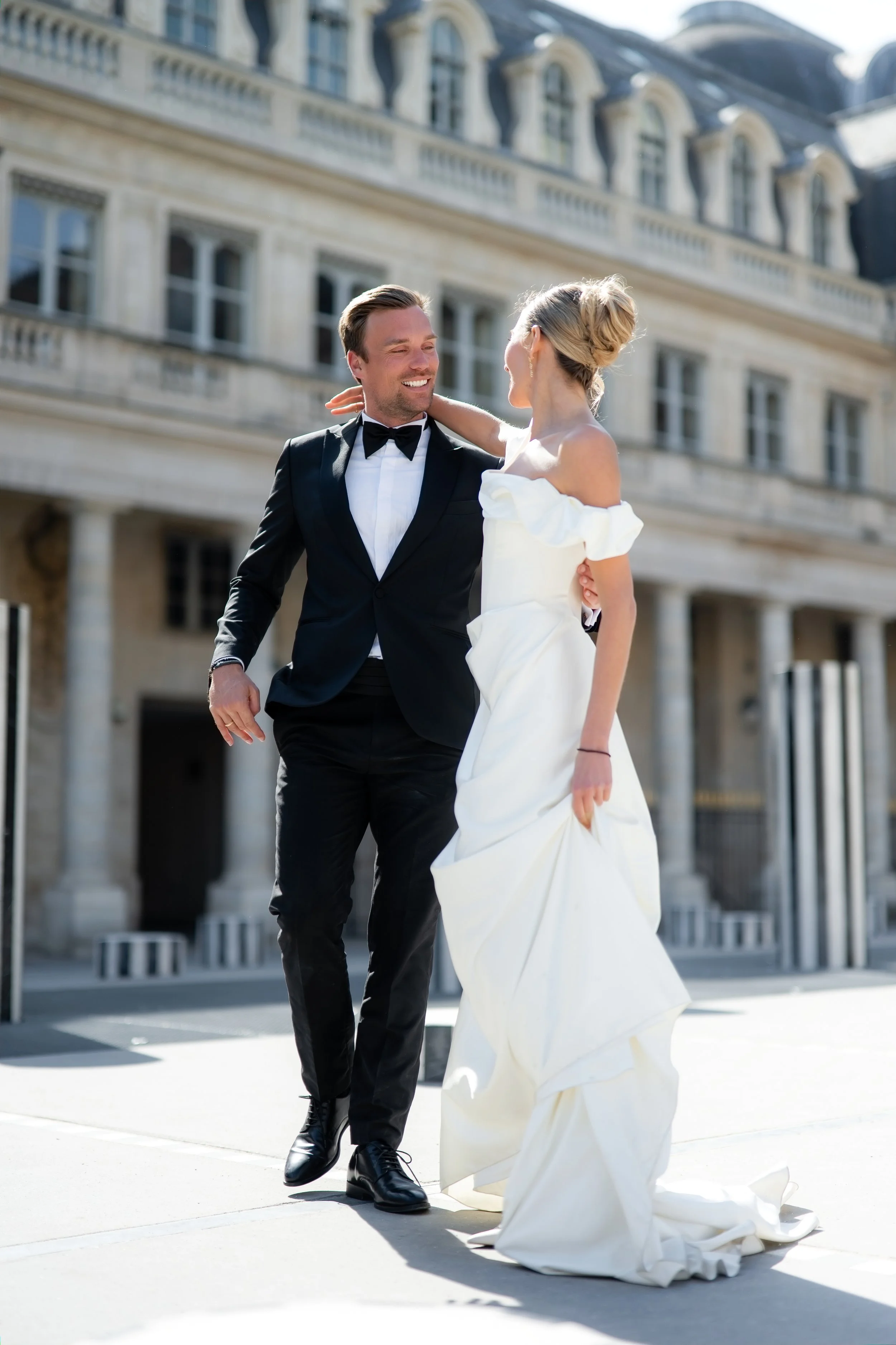 A man in a tuxedo and a woman in a white wedding dress dancing outdoors in front of a European-style building.