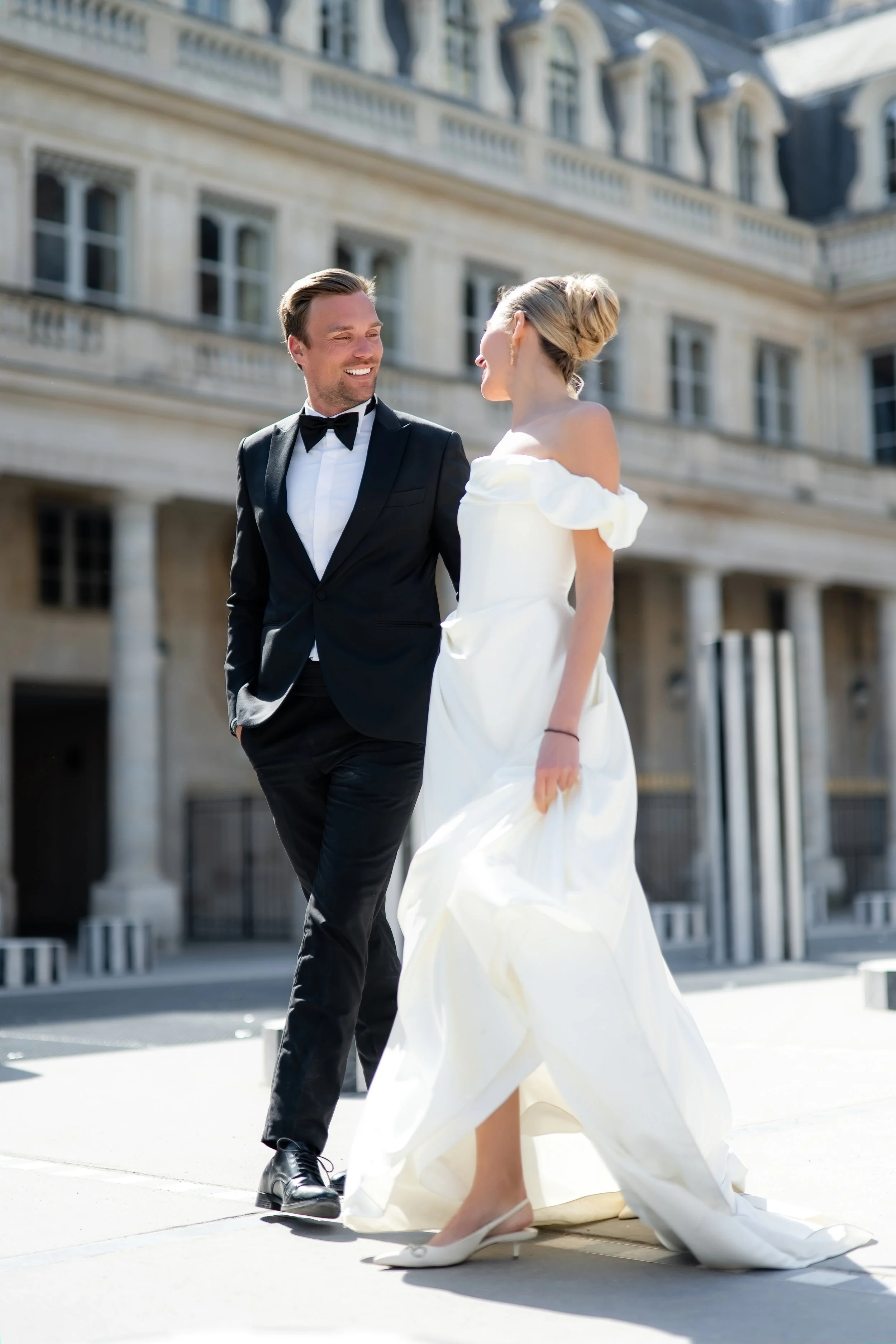 A man and woman in wedding attire standing outside in front of a historic building with classical architecture, smiling and looking at each other.