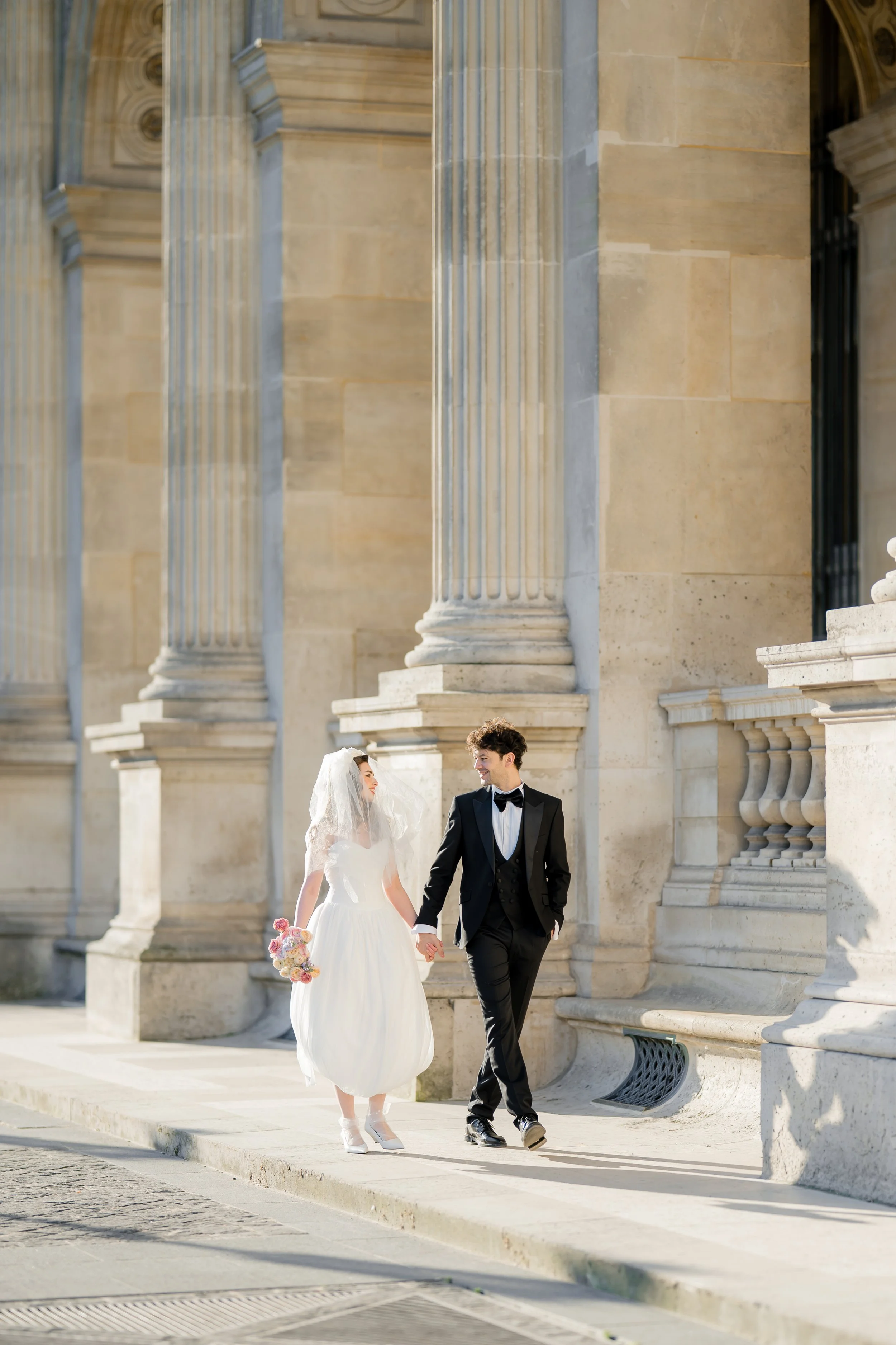 A bride and groom holding hands and walking in front of a historic building with large stone columns.