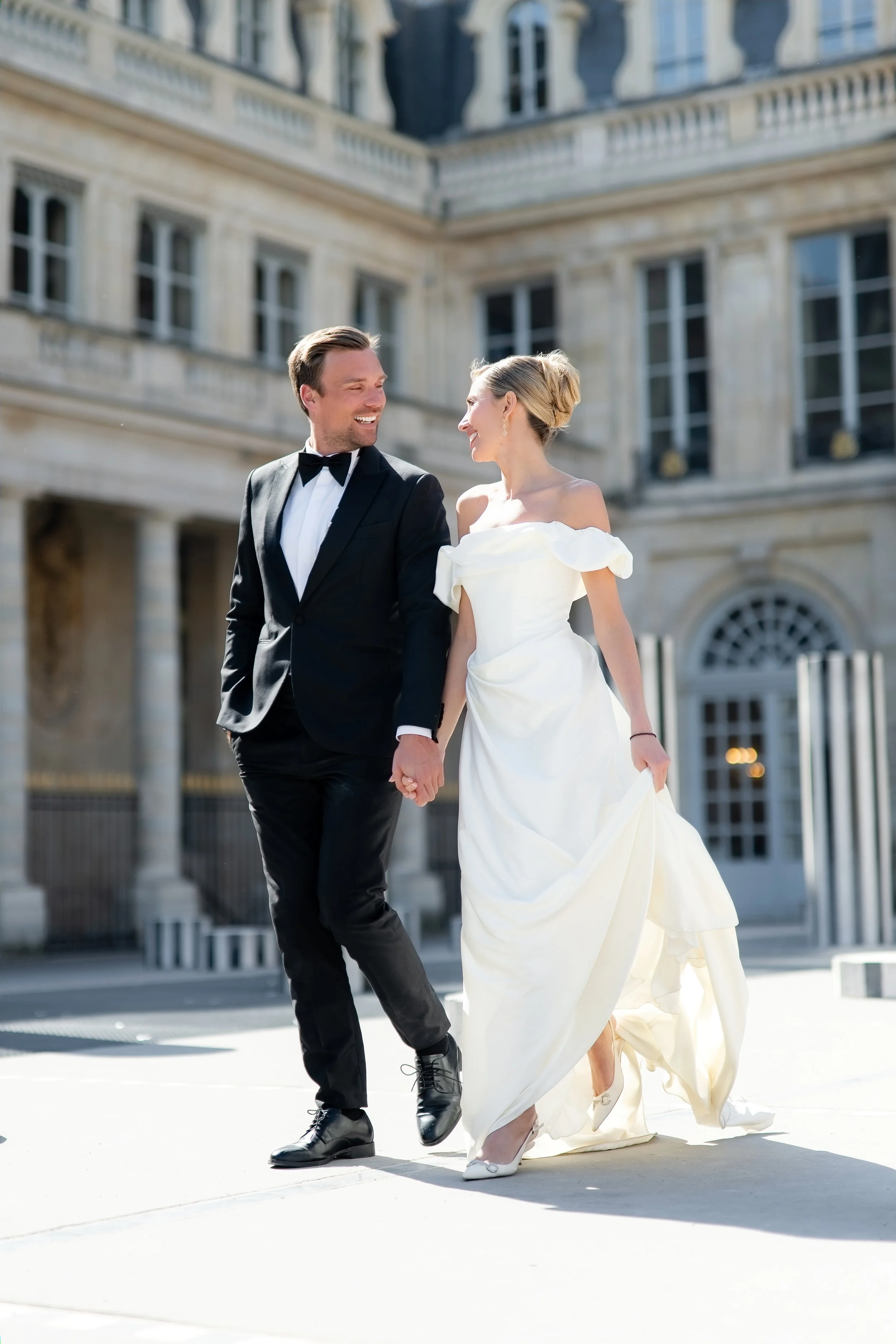 A couple dressed in formal wedding attire walking hand in hand outside a building with classic architecture; the groom is in a black tuxedo and the bride is in a white off-the-shoulder wedding gown.