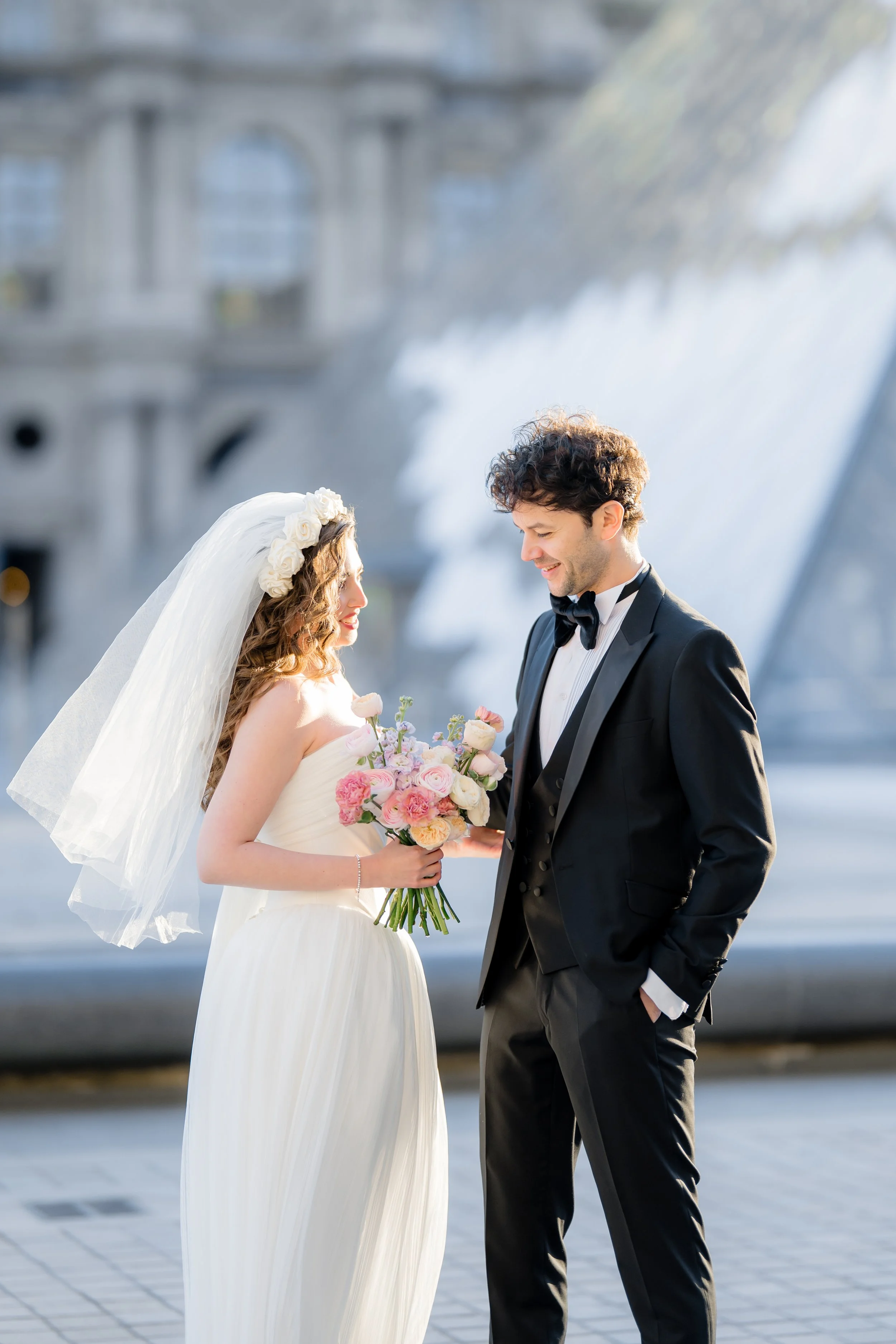 A bride wearing a white wedding dress with a veil and floral headpiece, holding a bouquet of pink and white flowers, smiles at a groom dressed in a black tuxedo with a bow tie, outside the Louvre