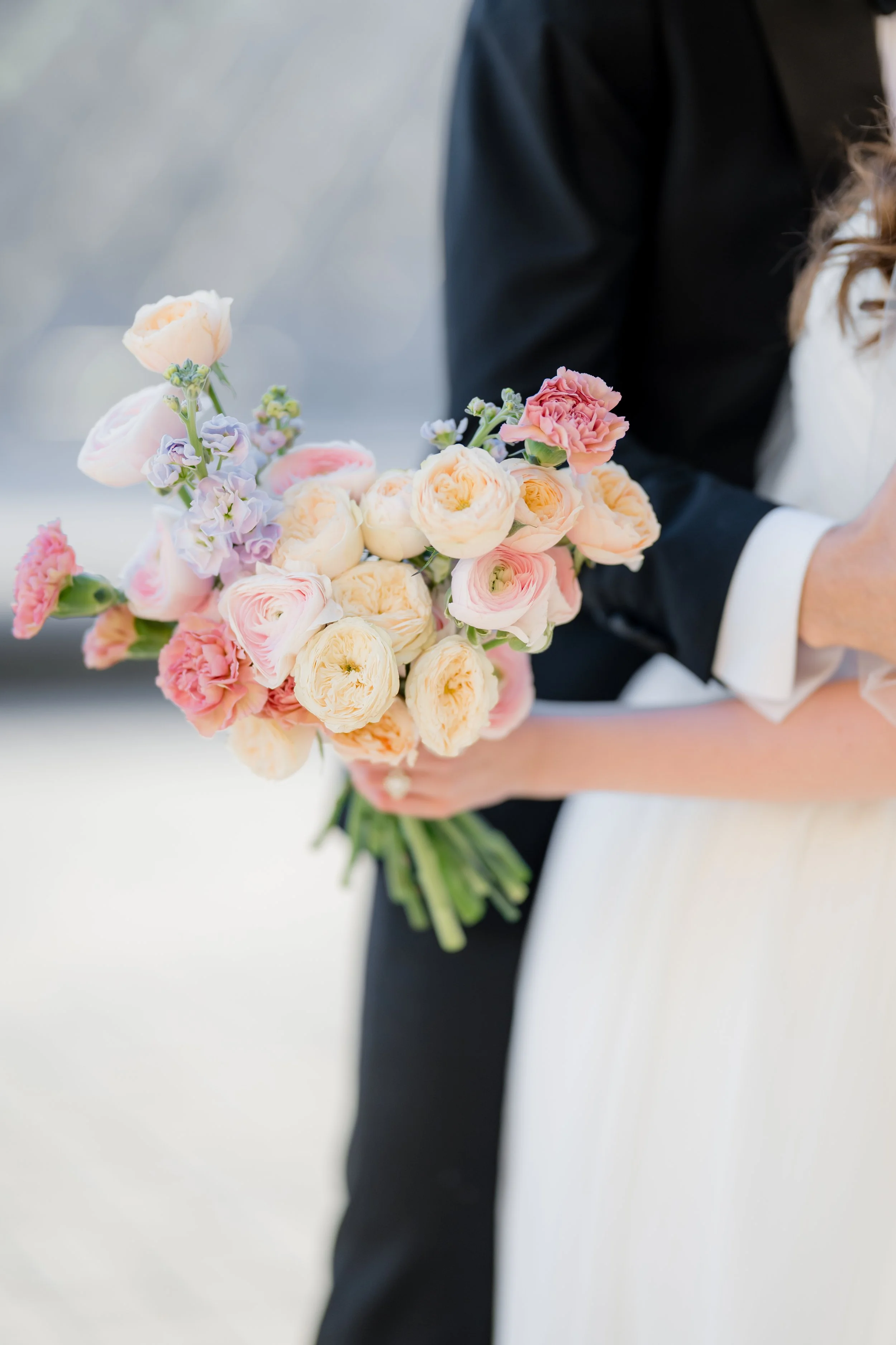 A person in a black suit holding a bouquet of pink, cream, and purple flowers.