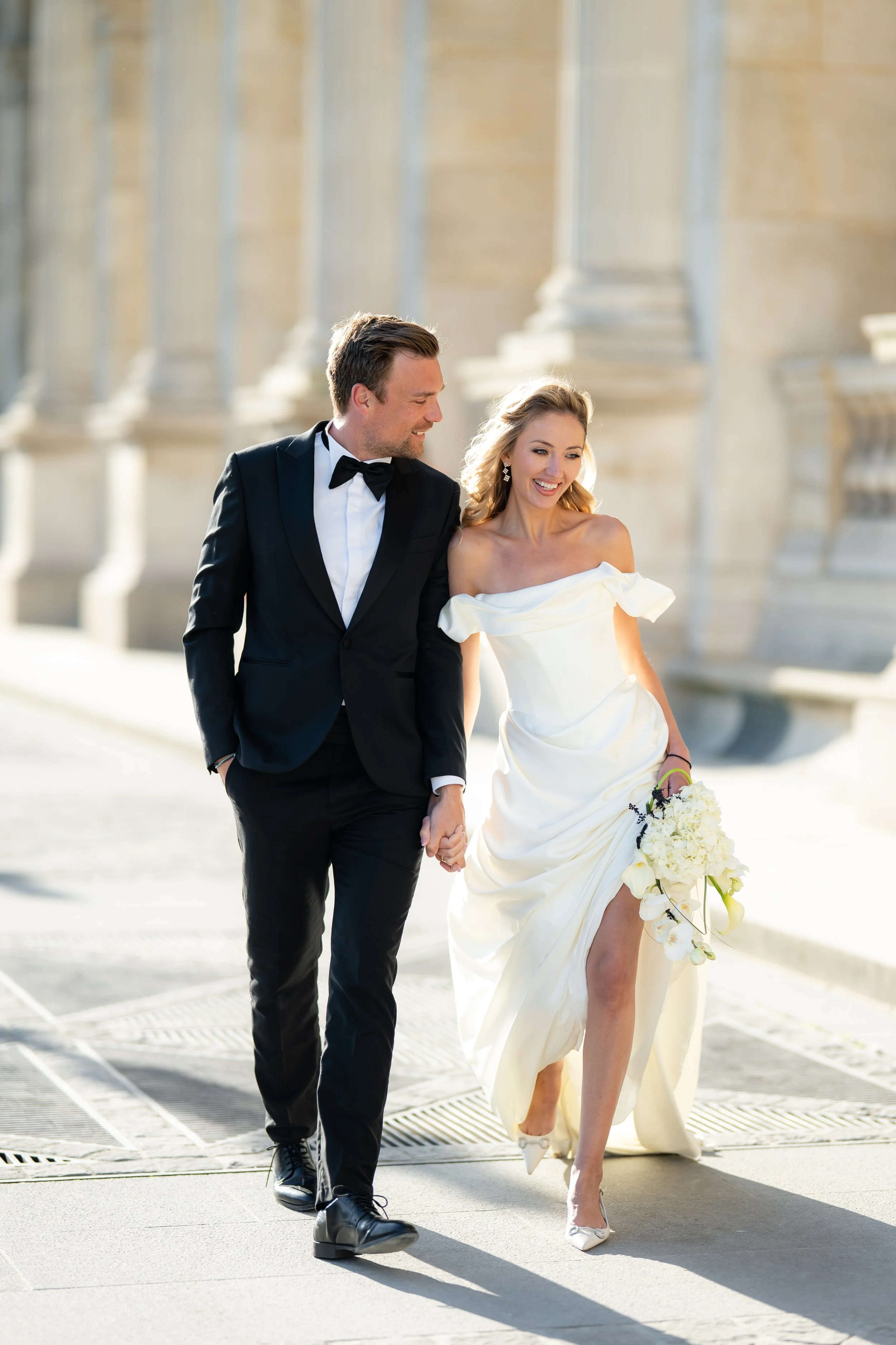 A newlywed couple walking outdoors, holding hands and smiling. The groom is wearing a black tuxedo with a bow tie, and the bride is in an off-shoulder white wedding gown holding a bouquet of white flowers. The background features large, classical arc