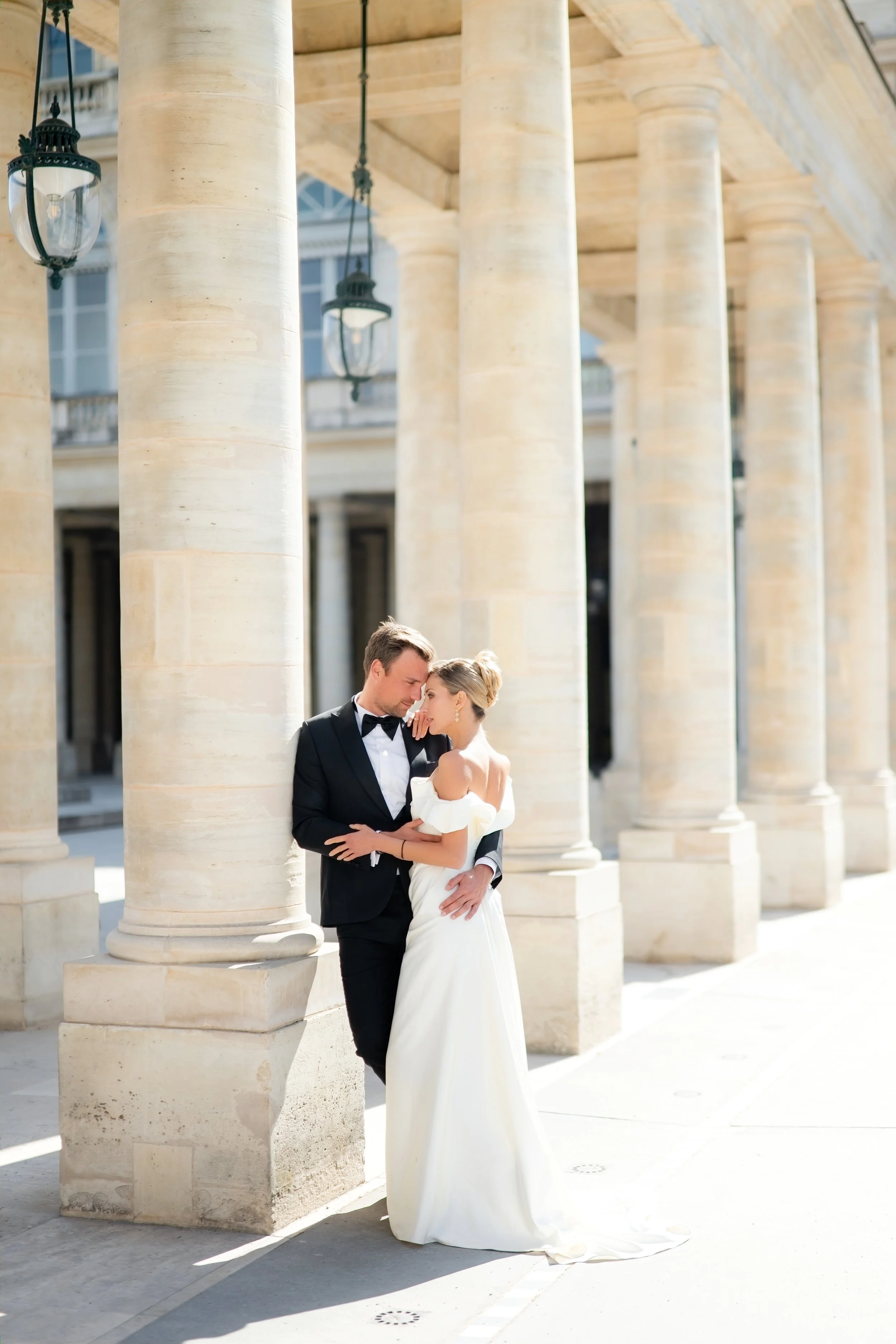 A newlywed couple dressed in wedding attire, standing close and sharing an intimate moment under classical stone columns with hanging street lamps, in front of a grand building.