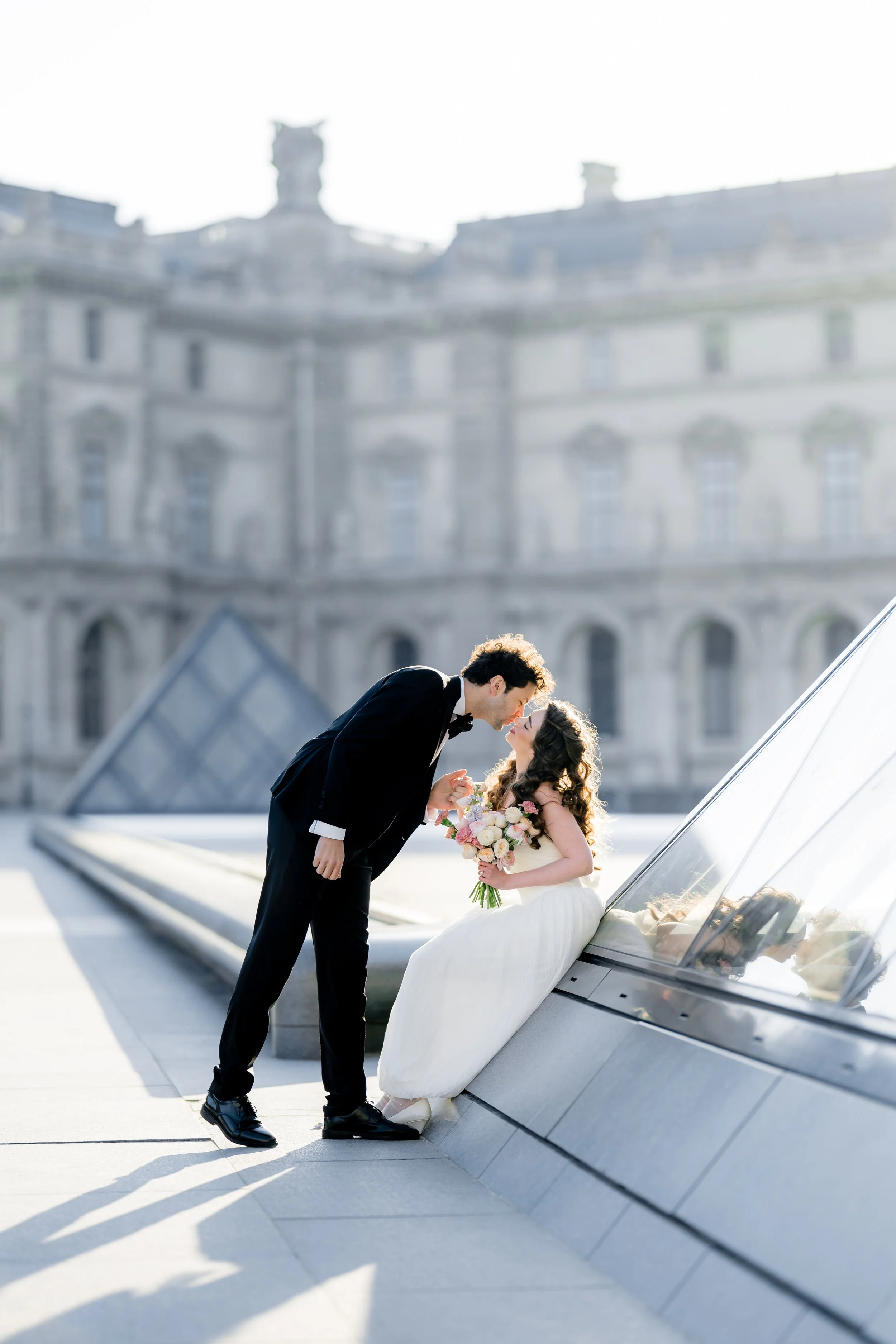 A bride and groom are in a romantic pose on a modern rooftop. The groom leans in towards the bride, who is sitting on a sleek white car. The bride holds a bouquet of flowers. The background features an elegant historic building with detailed architec