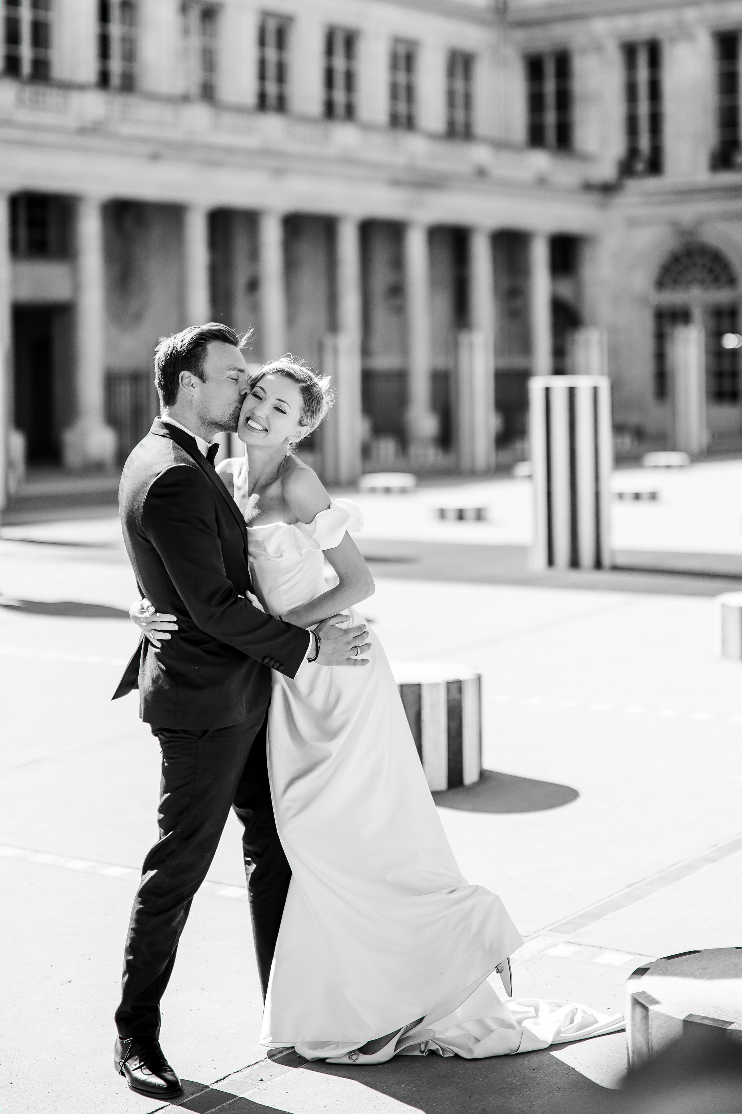 A bride and groom in wedding attire, smiling and embracing outdoors in front of a historic building with columns.