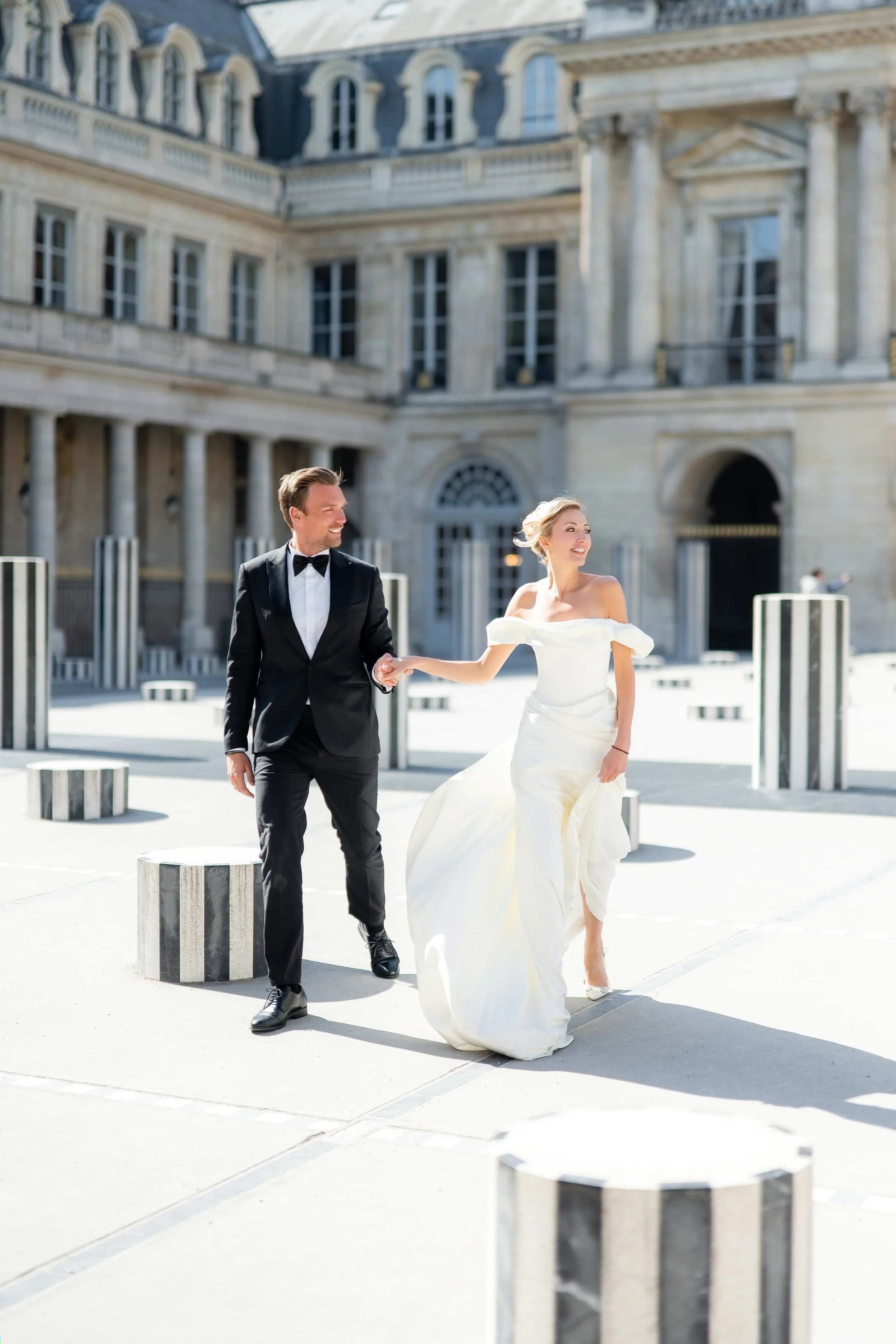 A bride and groom walking hand in hand outside a historic building, smiling and looking at each other, with the bride wearing a white off-shoulder gown and the groom in a black tuxedo.