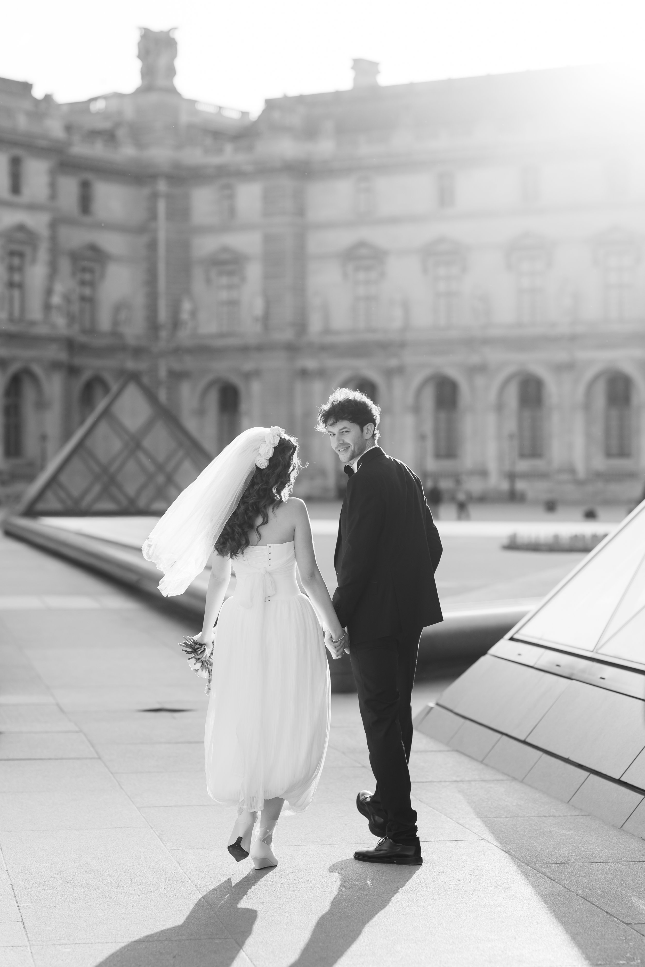 A bride and groom holding hands and smiling, with modern buildings in the background, during a wedding photoshoot in an outdoor urban setting.