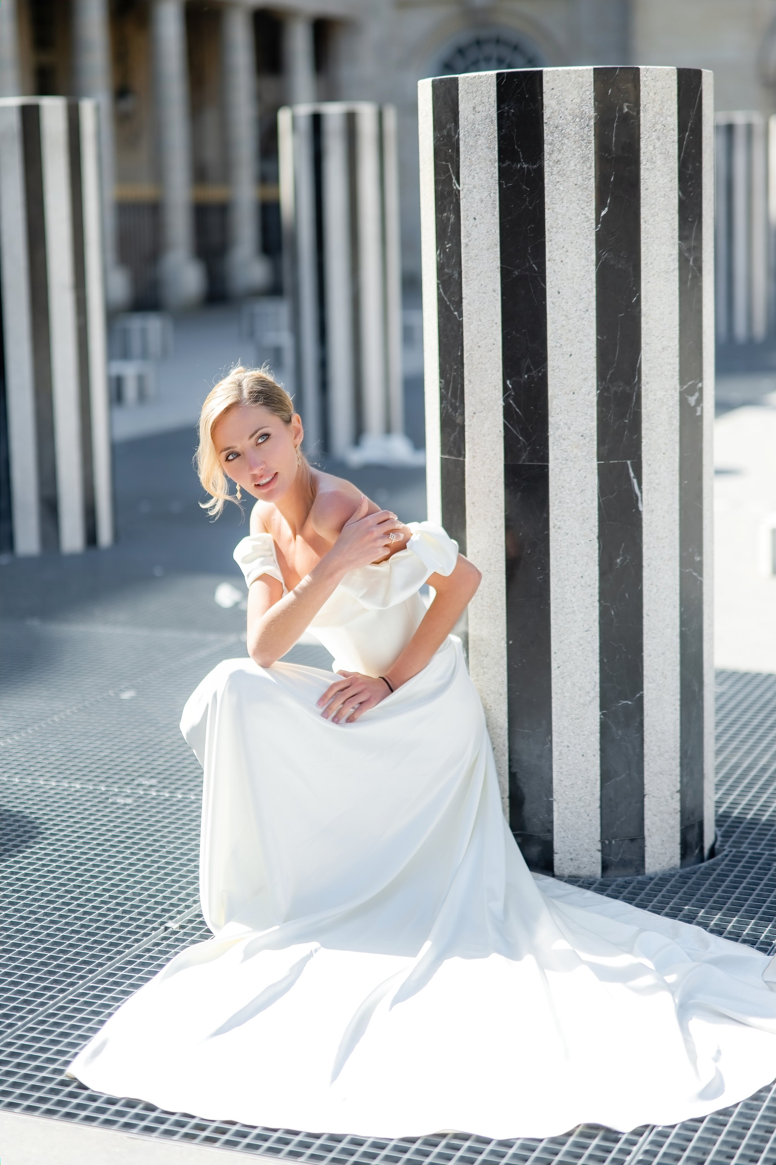 A woman in a white wedding dress sitting on a metal grate floor outdoors, with a large black and white striped column and blurred background of outdoor structures.