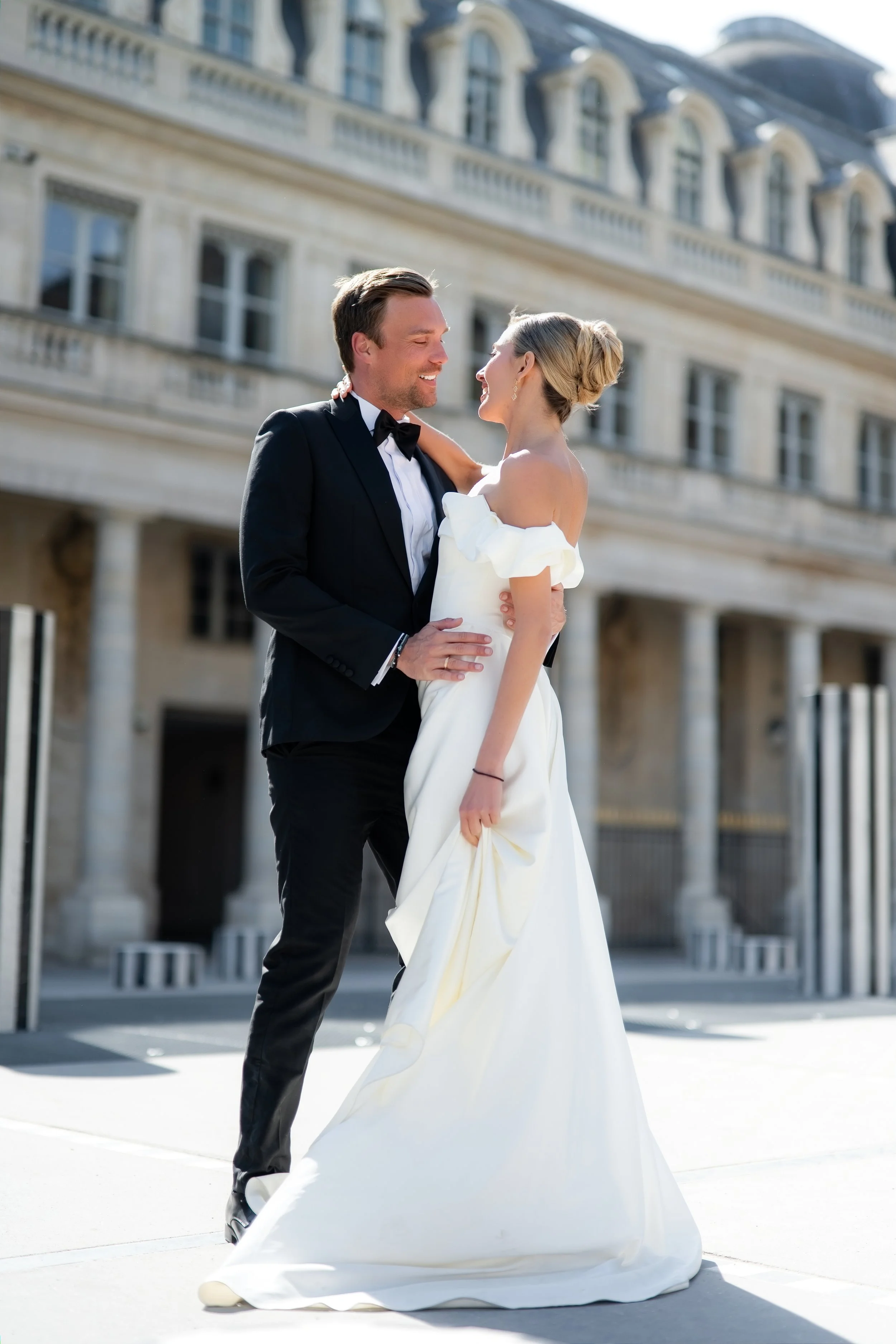 A man and woman in wedding attire holding each other and smiling outside a European-style building.
