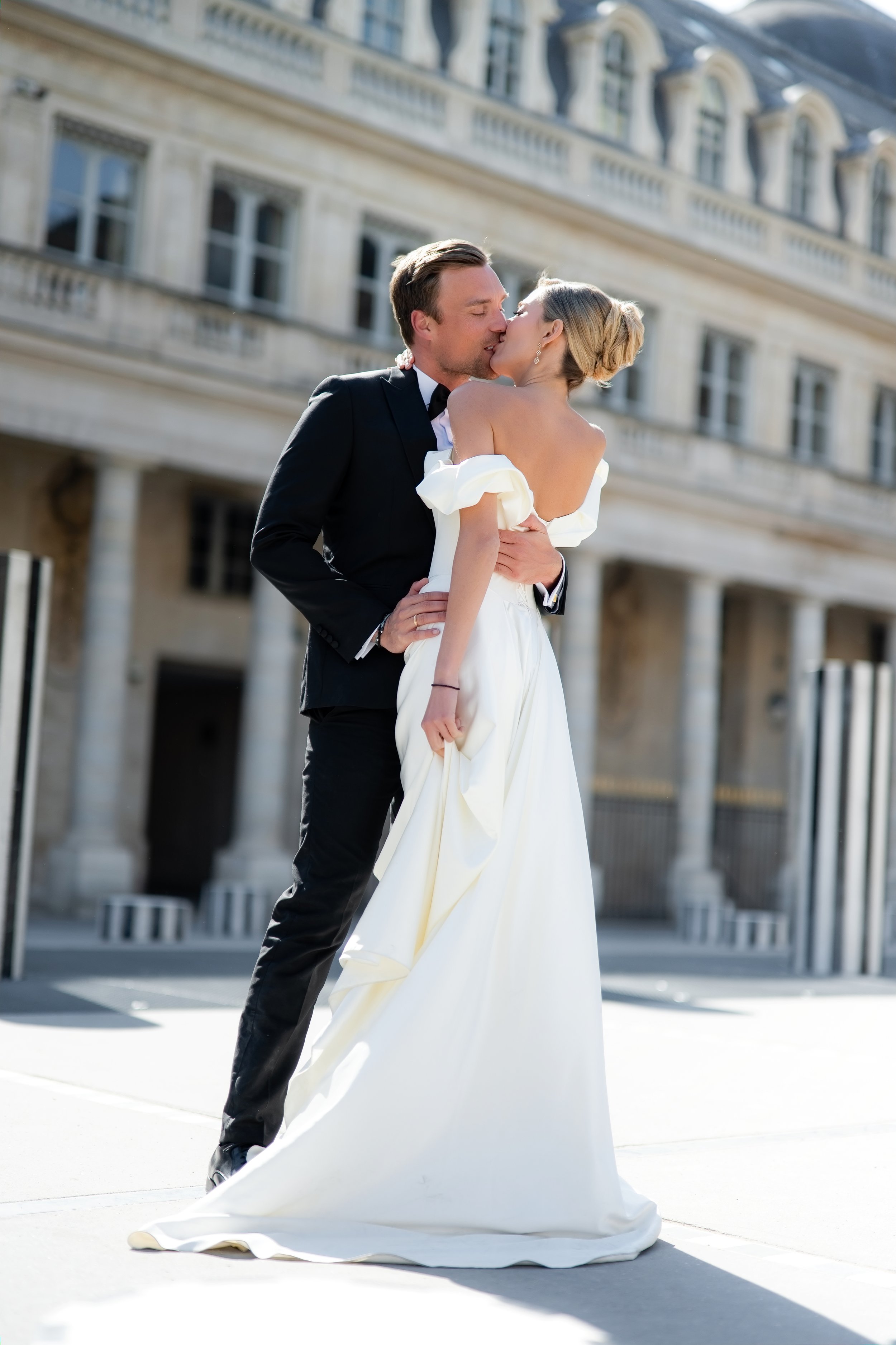 A newlywed couple sharing a kiss outdoors at Palais Royal, with the bride in a white wedding dress and the groom in a black tuxedo.
