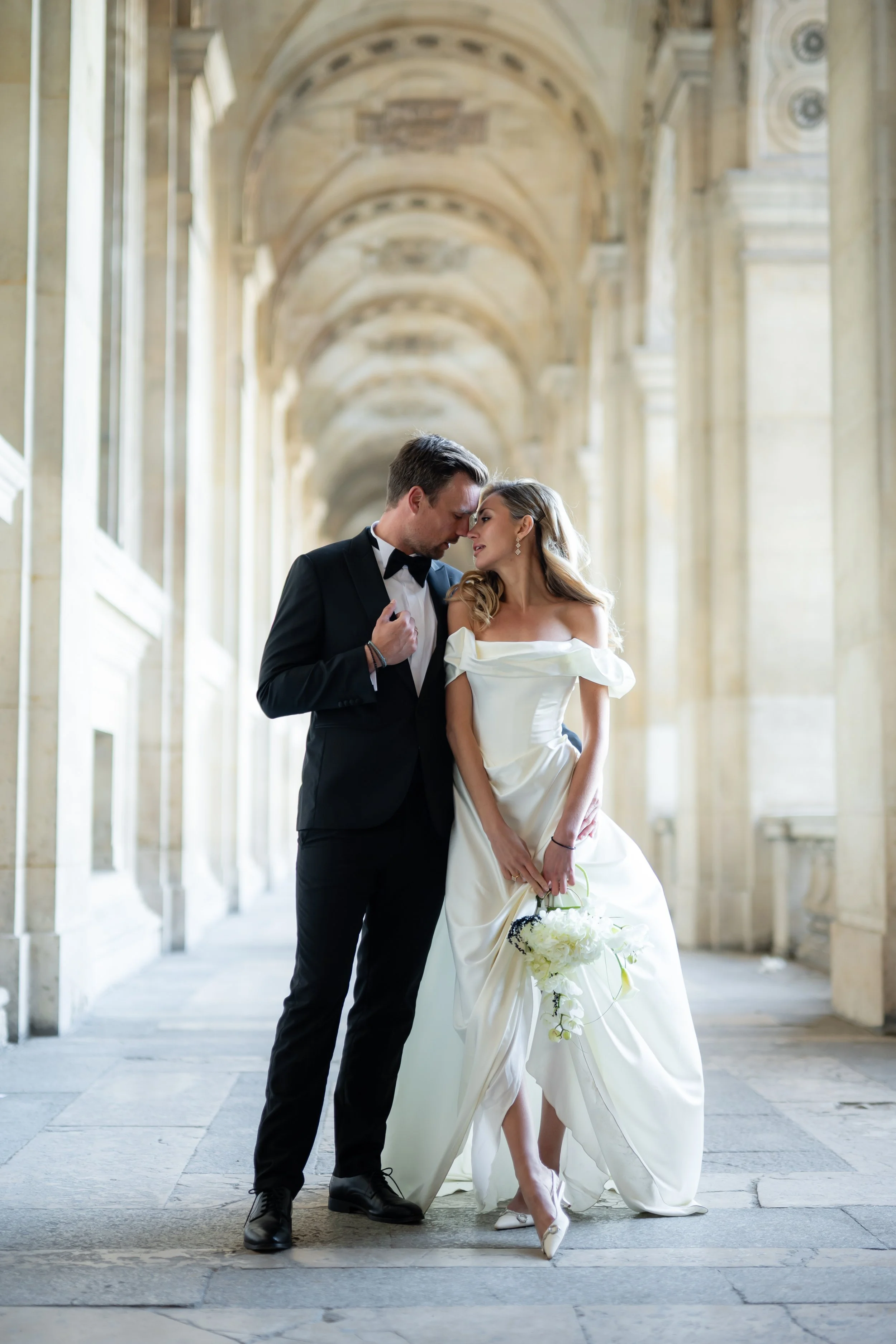 A groom and bride are posing closely in a grand hallway with high arched ceilings and stone columns. The groom is dressed in a black tuxedo and bow tie, and the bride is wearing an off-shoulder white wedding gown, holding a bouquet of white flowers. 