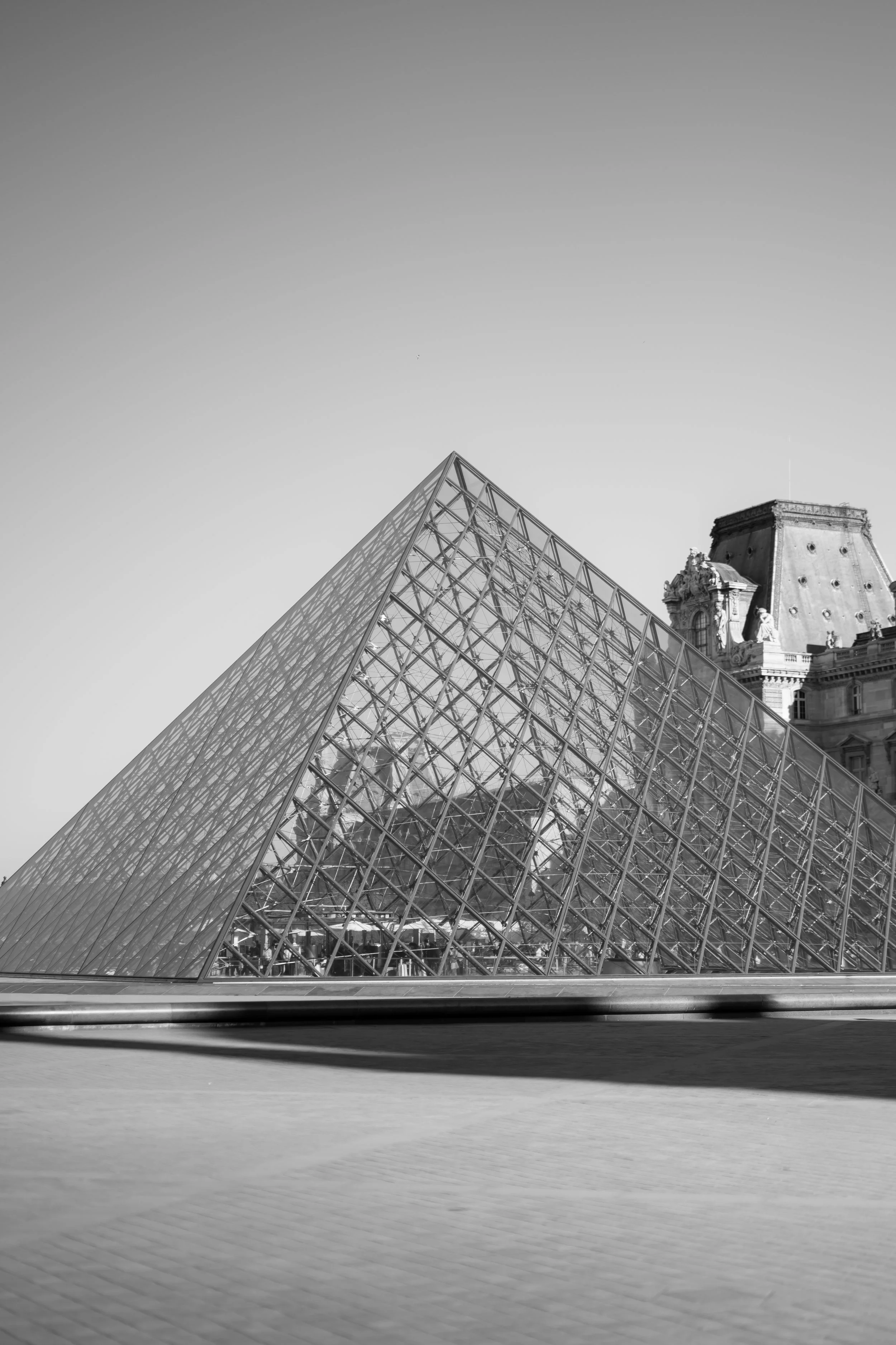 Black and white photo of the Louvre Pyramid in Paris, France, with parts of historic buildings visible in the background.