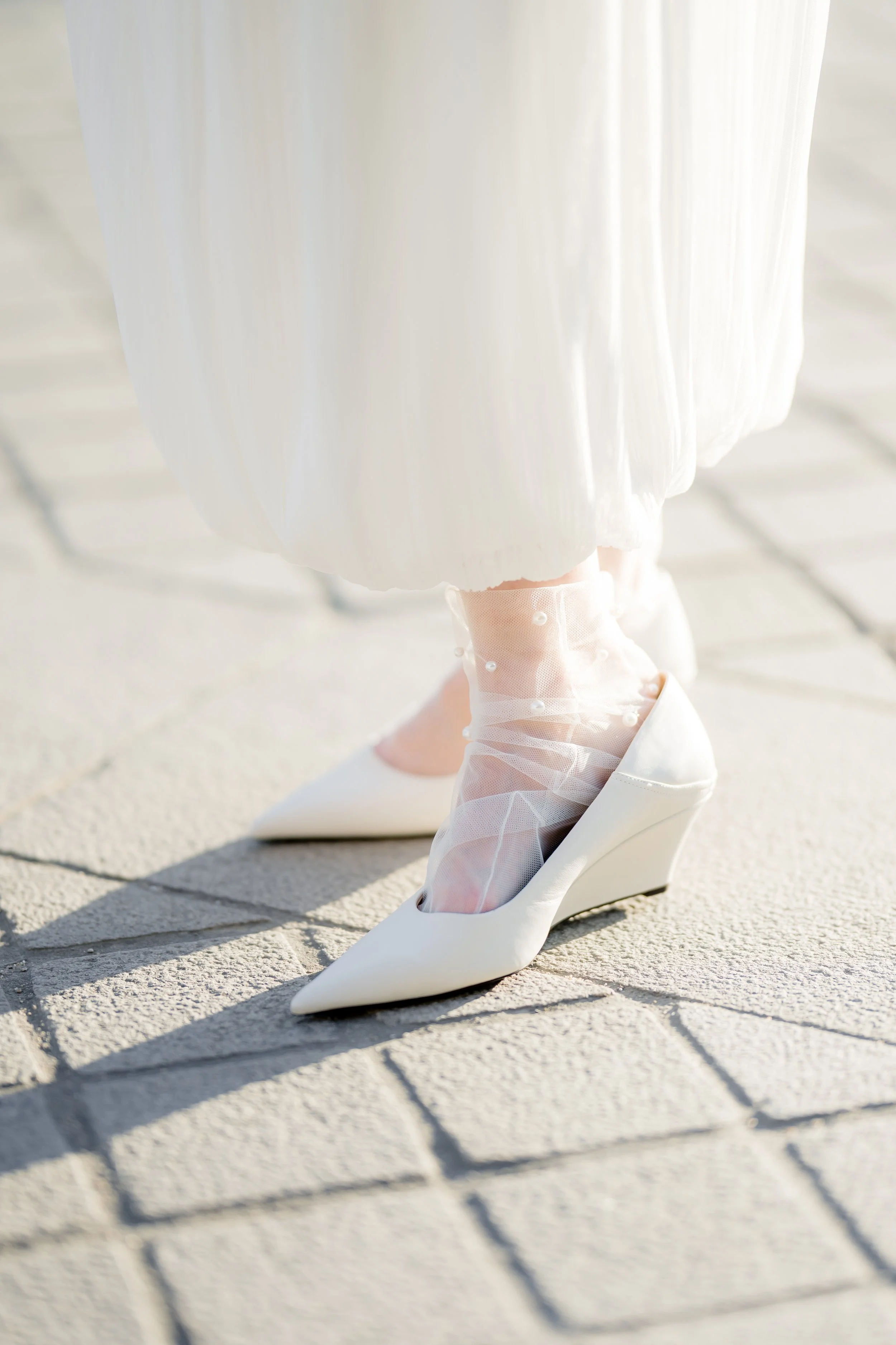 Close-up of a woman's feet in white high-heeled shoes, with lace stockings decorated with pearls, walking on a tiled sidewalk.