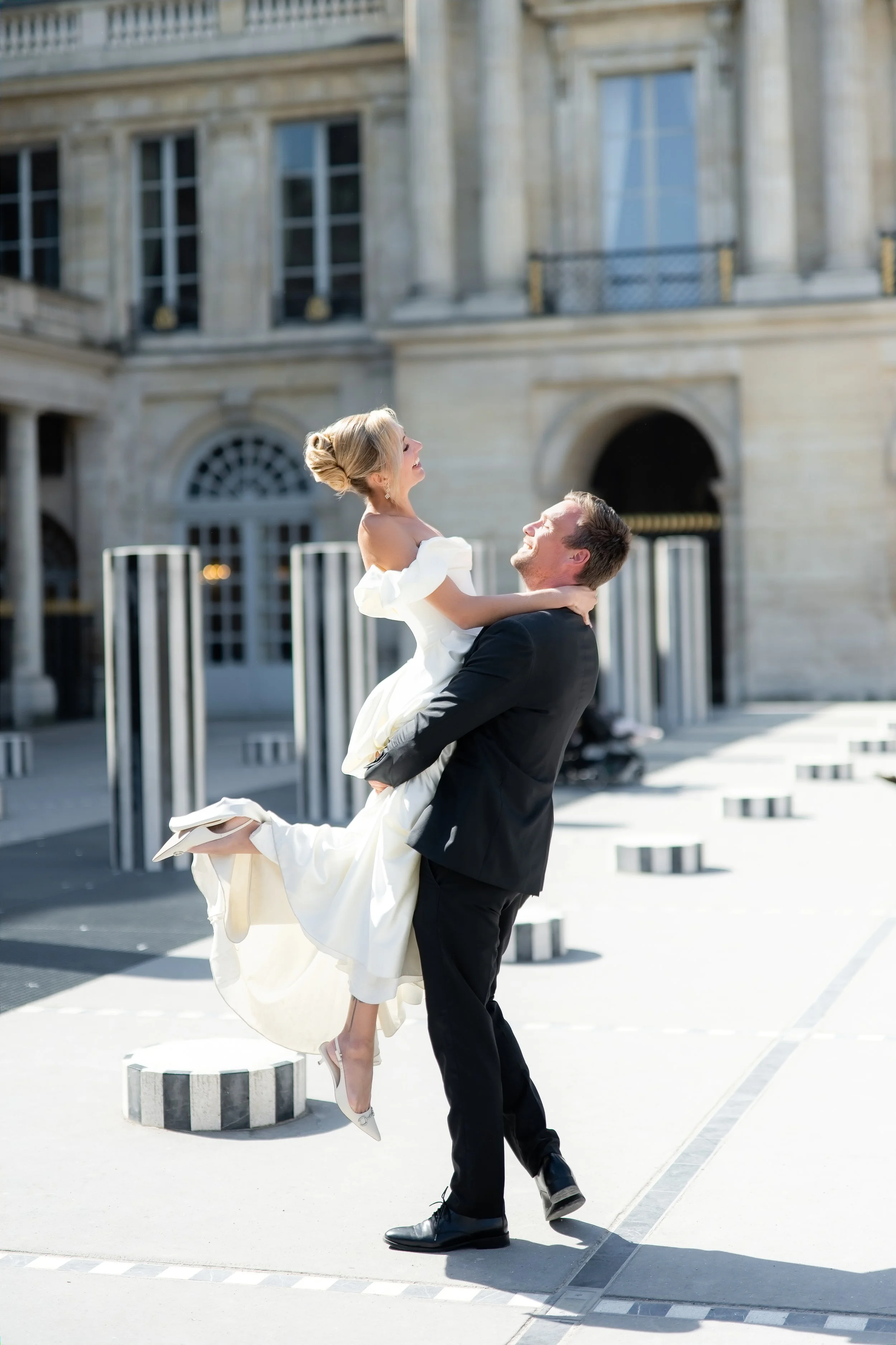 A man in a black suit is lifting a woman in a white dress in the air outdoors in front of a large building. The woman is smiling and looking upward, while the man is looking at her with a joyful expression.
