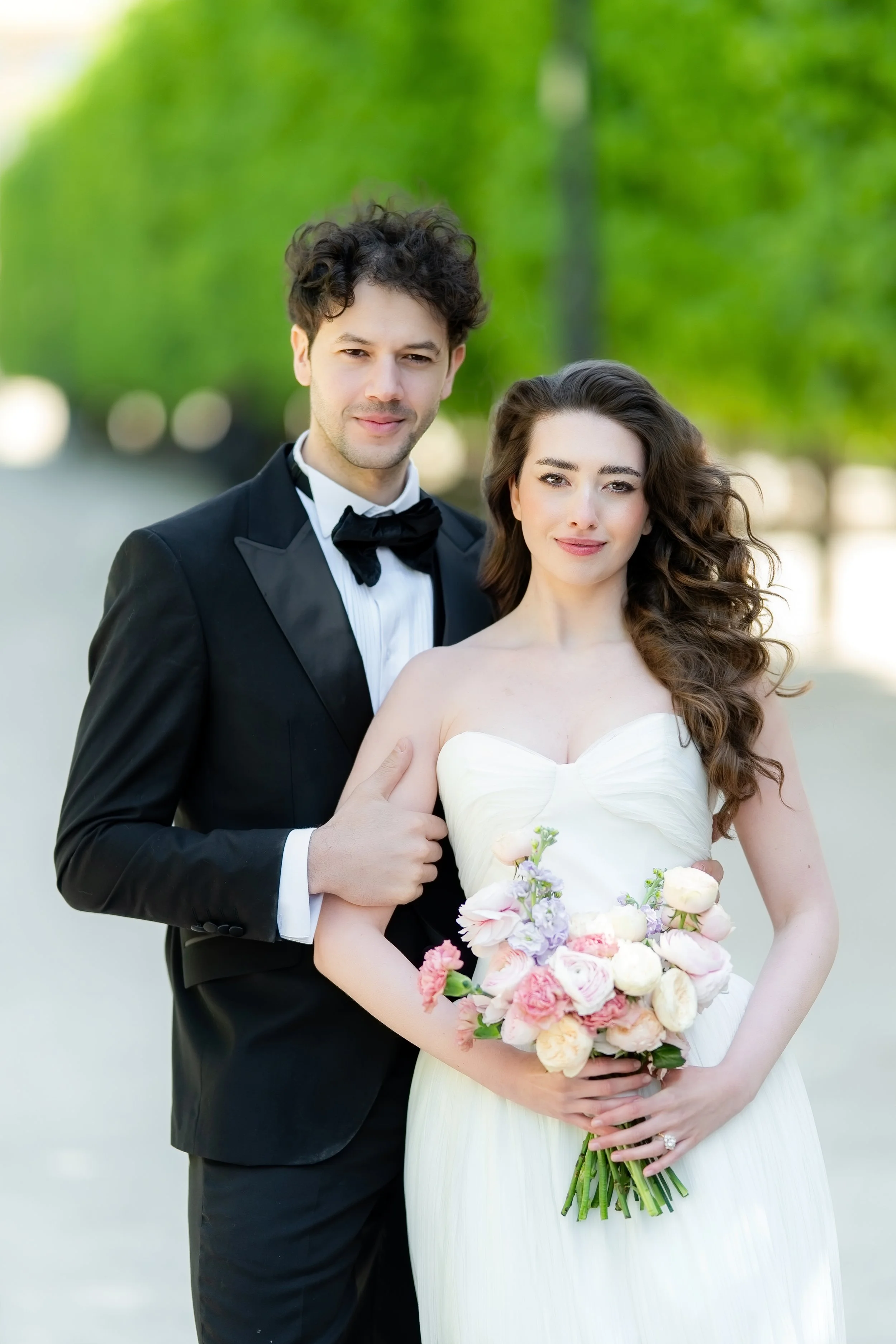 A newlywed couple standing outdoors with green trees in the background. The groom is wearing a black tuxedo with a bow tie, and the bride is wearing a white wedding dress and holding a bouquet of various colored flowers.