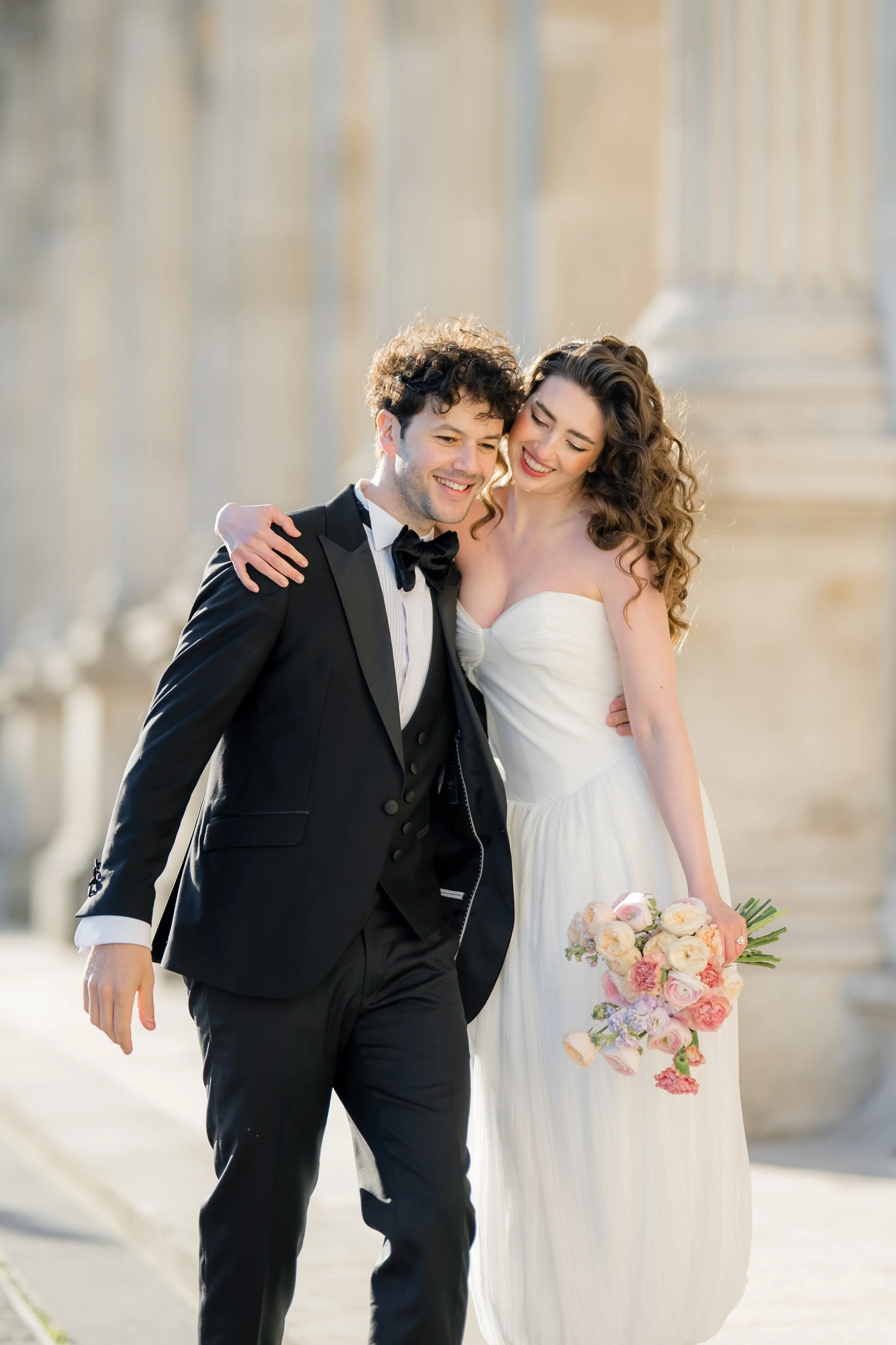 A newlywed couple walking outdoors near columns, smiling and enjoying each other's company. The groom is wearing a black tuxedo with a bow tie, and the bride is in a white wedding gown holding a bouquet of pink and white flowers.