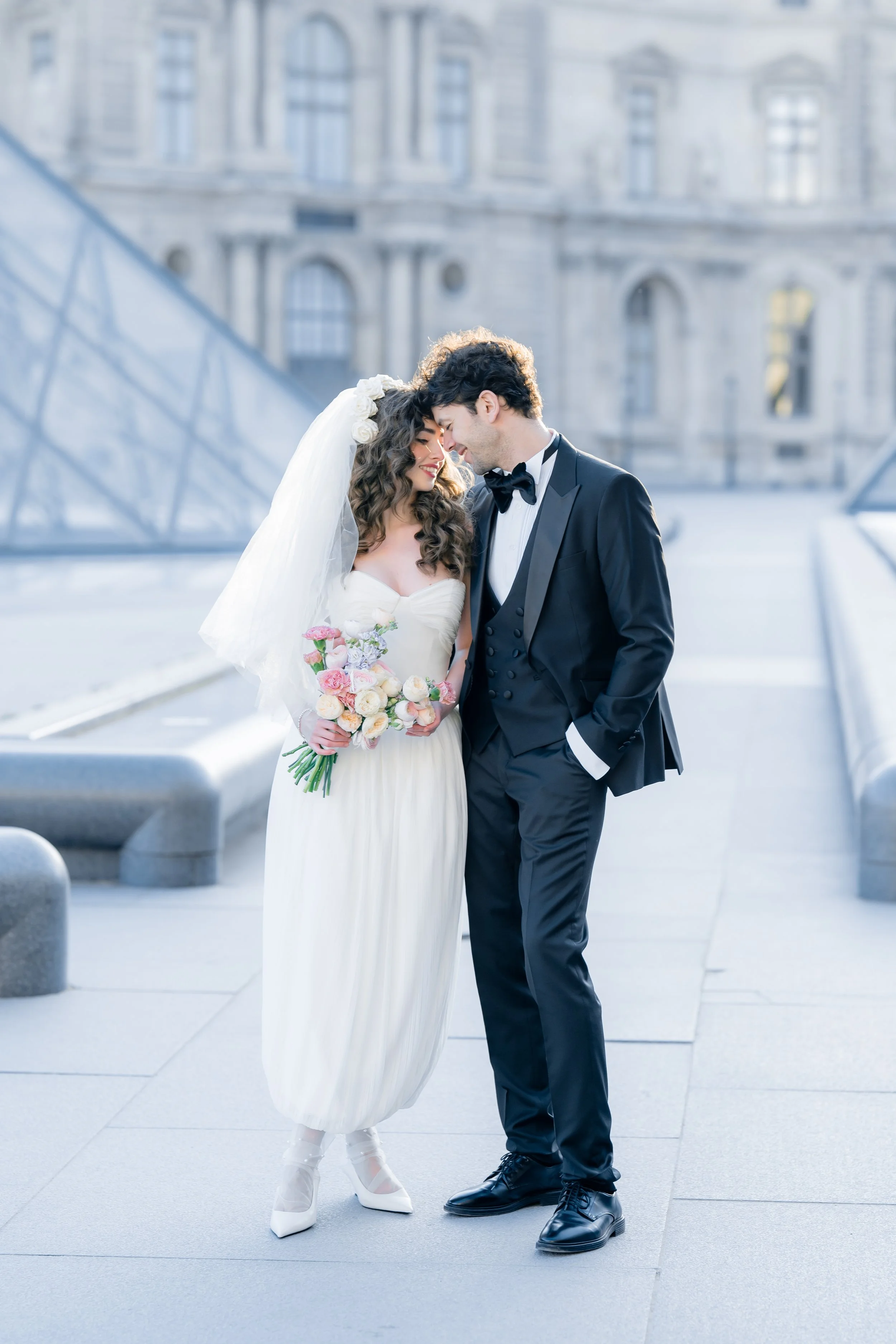 A bride and groom smiling and touching foreheads outdoors in of the louvre, on a sunny day. The bride is in a white gown holding a bouquet, and the groom is in a black tuxedo with a bow tie.