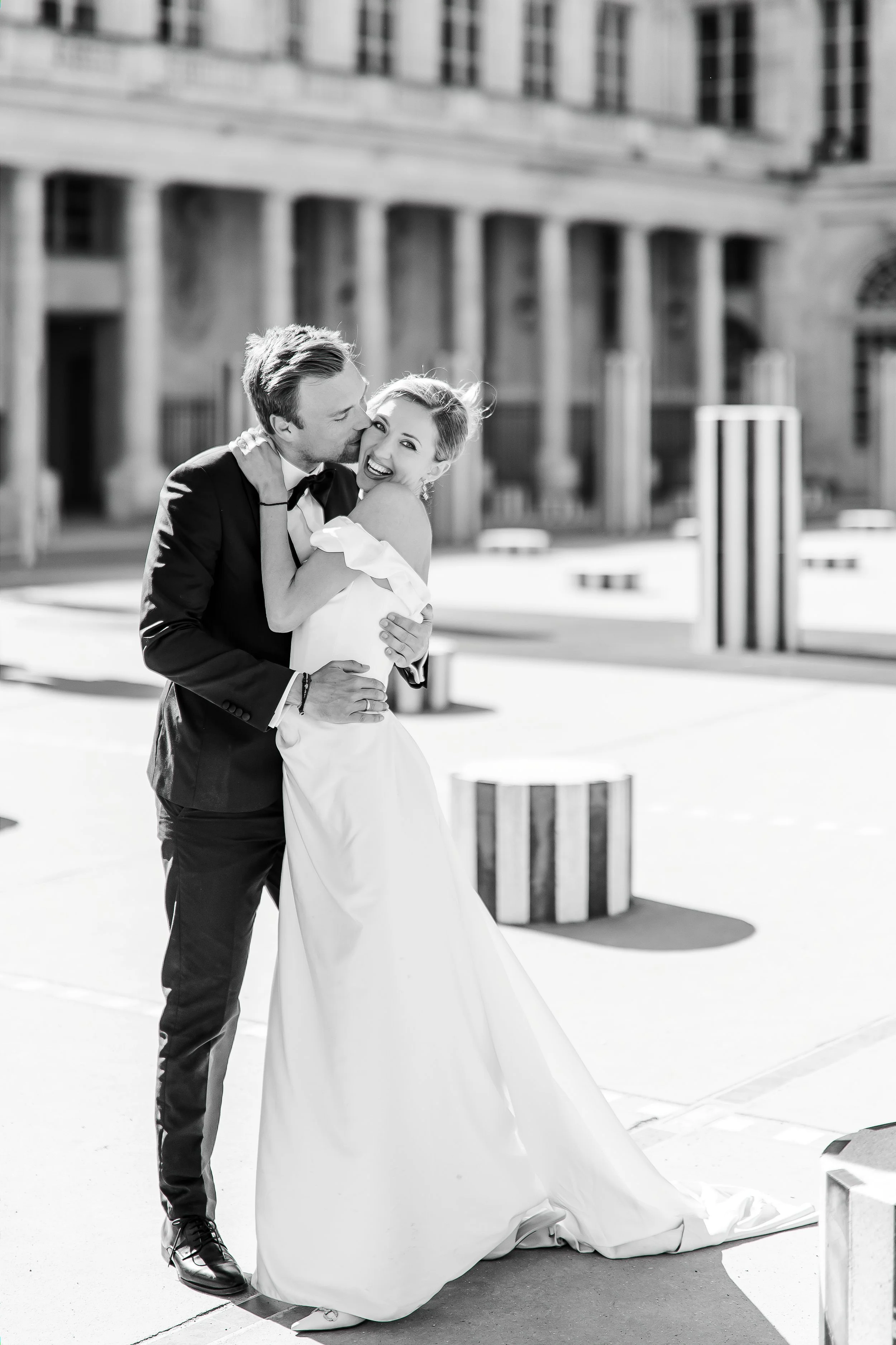 A black-and-white photo of a bride and groom embracing each other outdoors near a building with large columns. The groom is wearing a tuxedo and the bride is in a wedding dress, smiling happily.