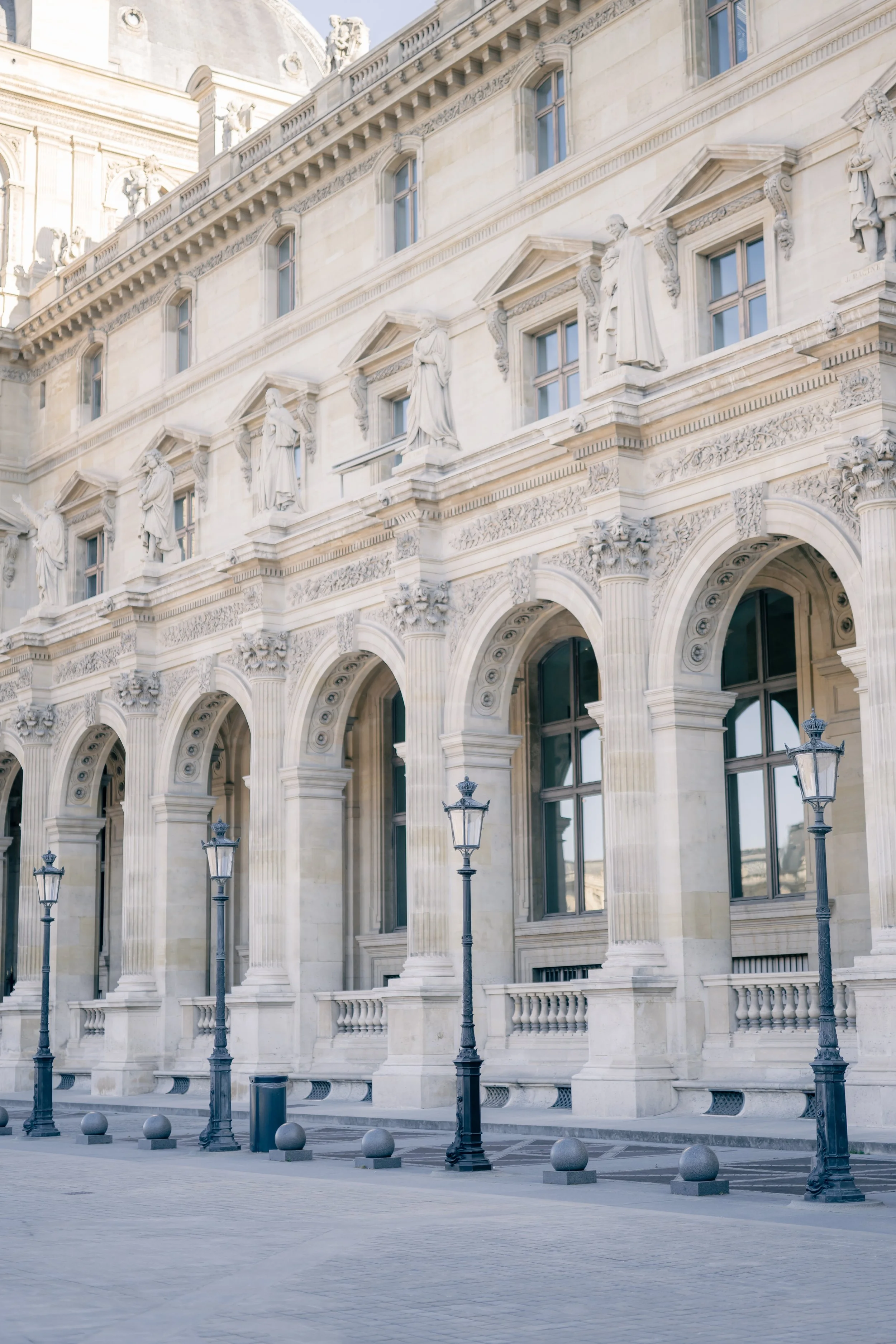 Historic classical building with ornate statues on the facade, large arched windows, and decorative architectural details. Street lamps and bollards in front.