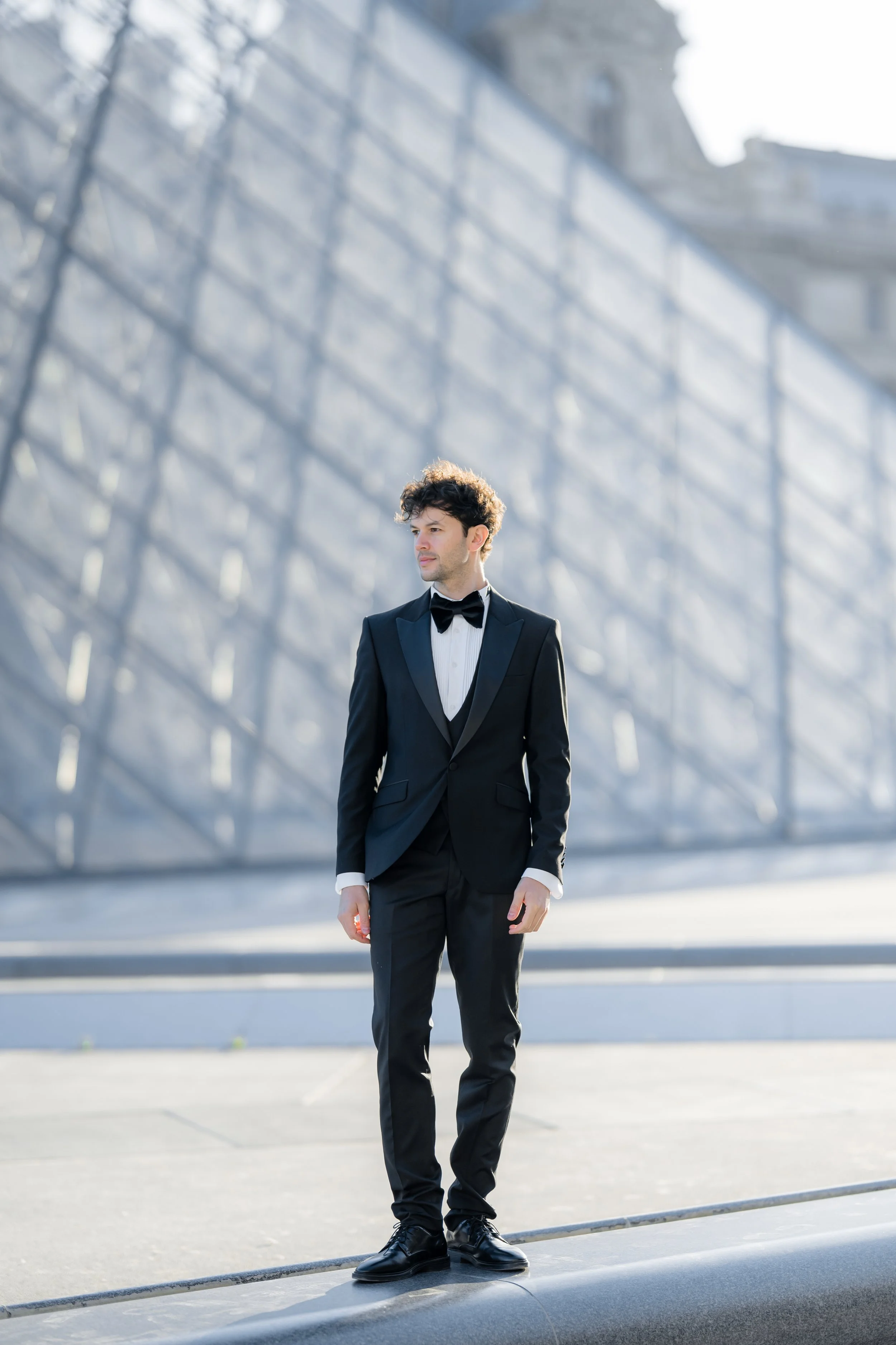 A young man in a black tuxedo with a black bow tie and white shirt stands outdoors on a city street. Modern glass and steel buildings are in the background, with sunlight casting shadows.
