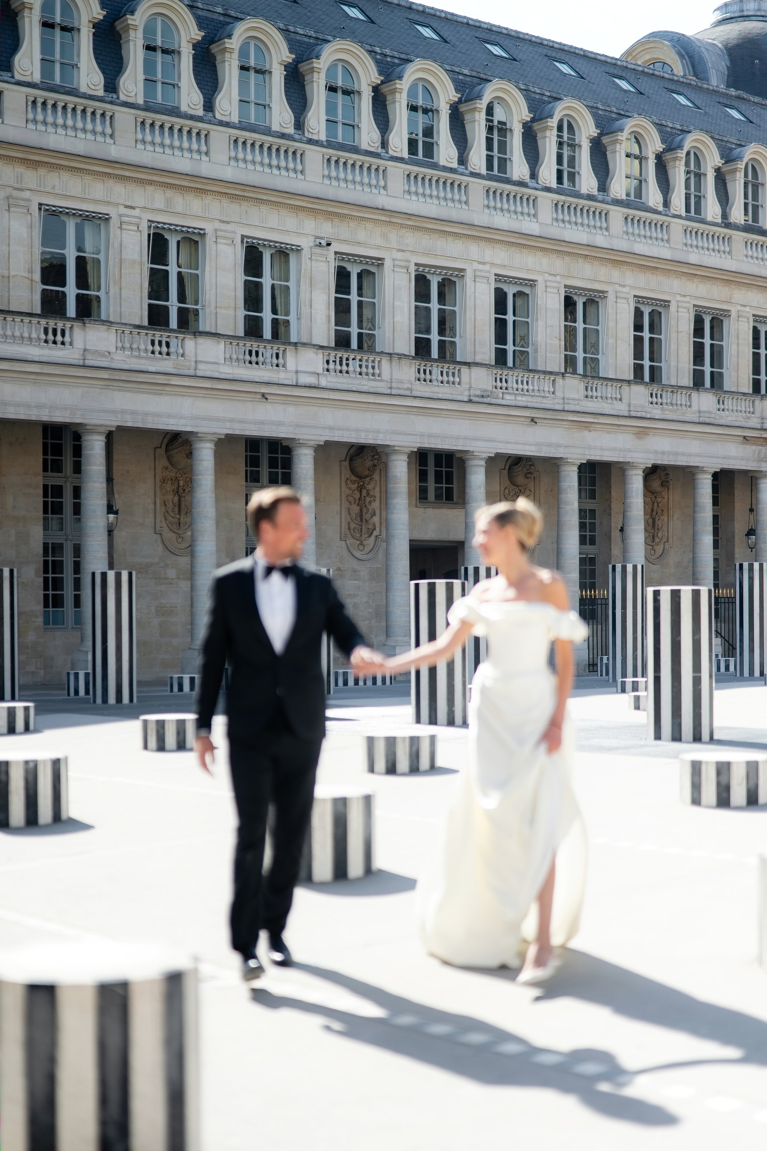 A couple dressed in formal wedding attire holding hands, walking outdoors in front of a grand building with classical architectural features, including multiple stories, large windows, and decorative columns.