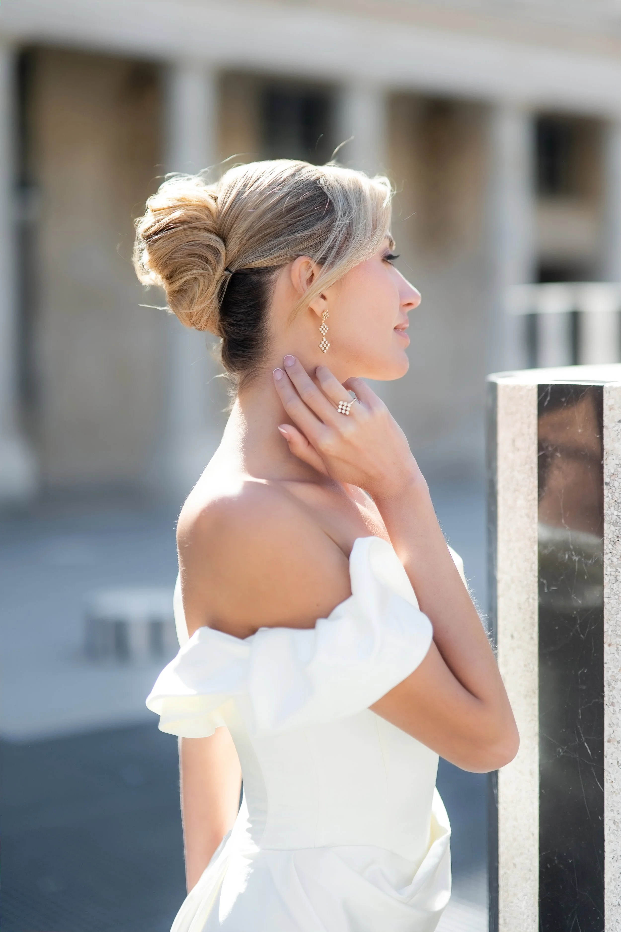 A woman with blonde hair styled in an elegant updo, wearing pearl earrings and a white off-shoulder dress, is touching her neck and standing outdoors near a marble railing with a blurred building in the background.